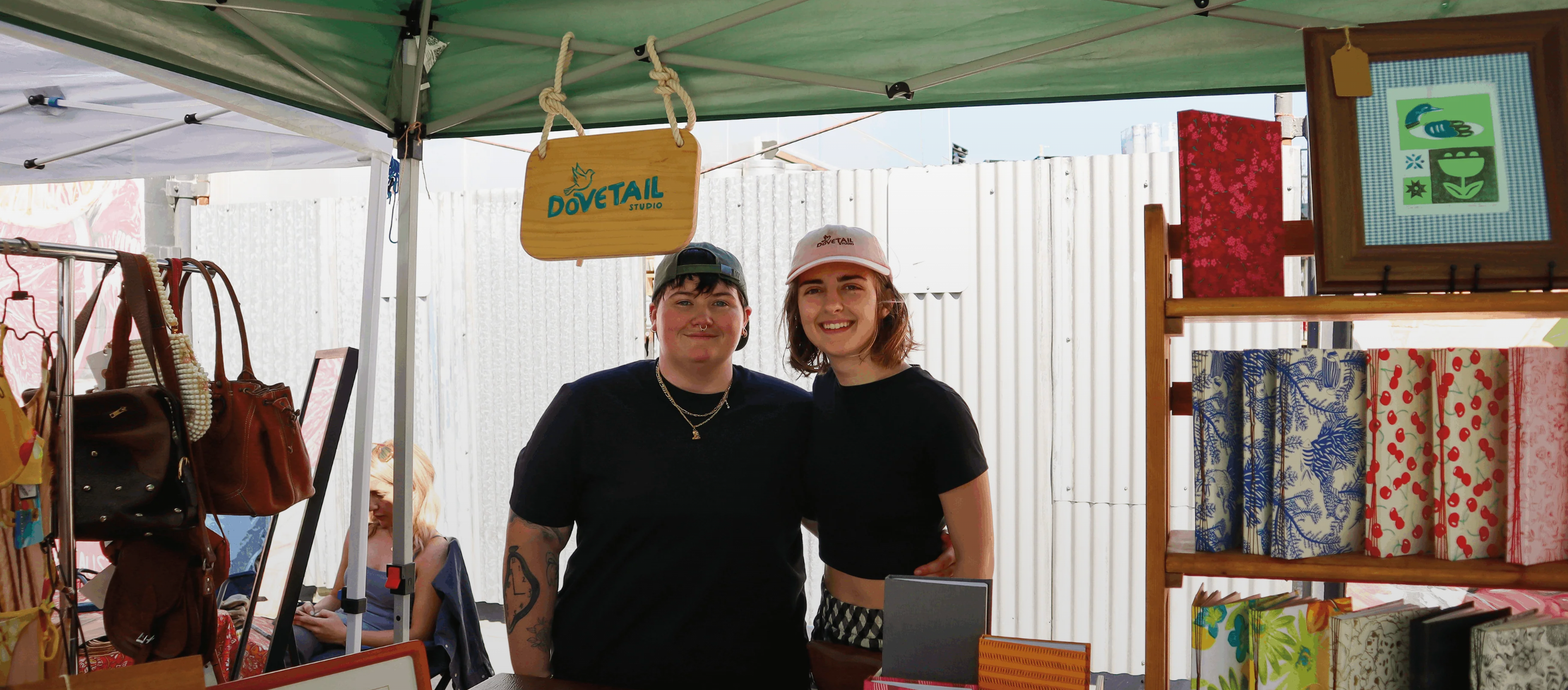 picture of the owners of dovetail studio Lilli Jones and Bella Thompson standing in their pop-up market setup with handbound journals, framed lonocut prints, a wooden sign, and stationery in the background. 