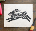 A black ink linocut print of a leaping hare on textured off-white handmade paper, displayed flat on a scratched dark wood surface with art tools partially visible around the edges.