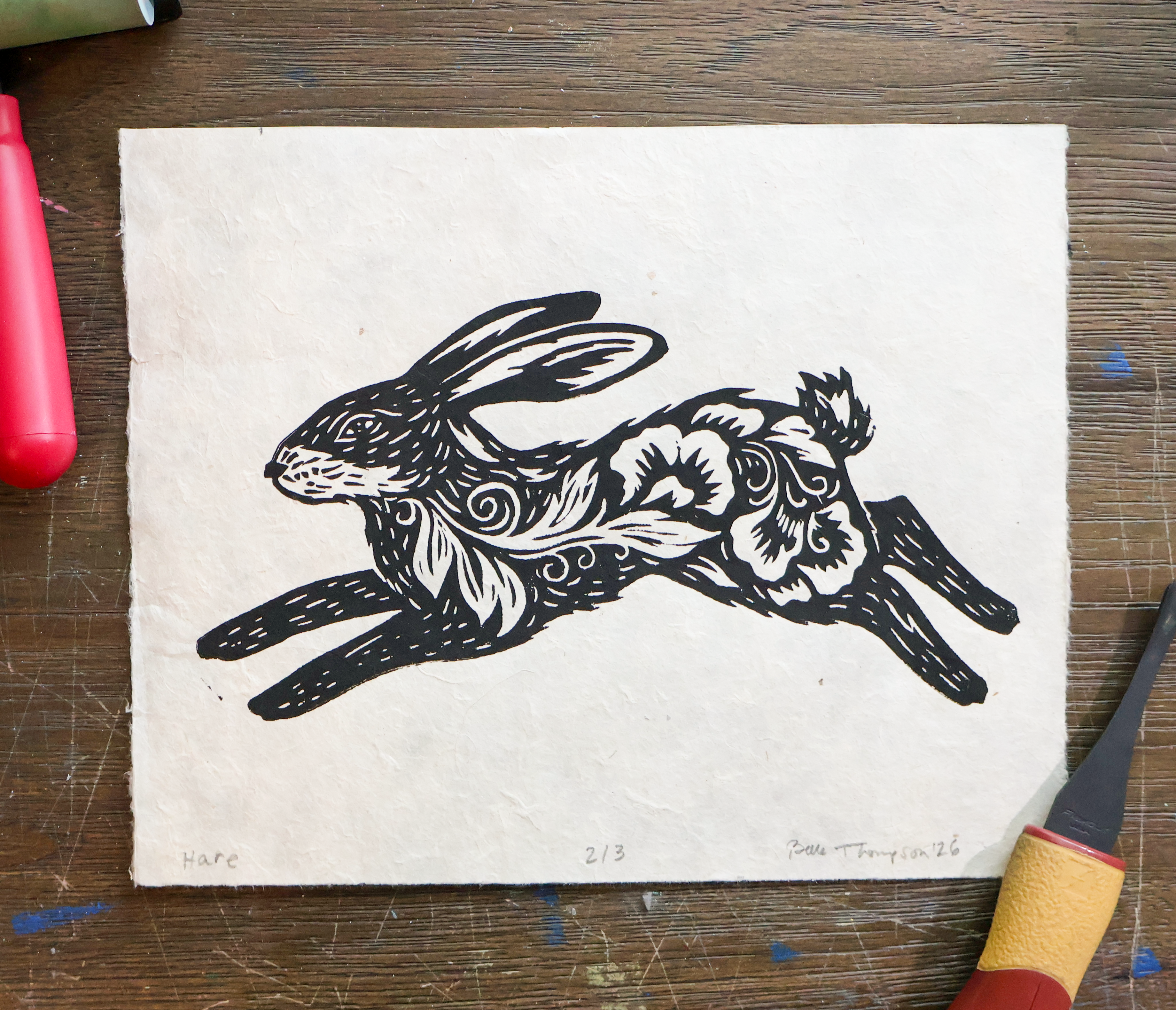 A black ink linocut print of a leaping hare on textured off-white handmade paper, displayed flat on a scratched dark wood surface with art tools partially visible around the edges.