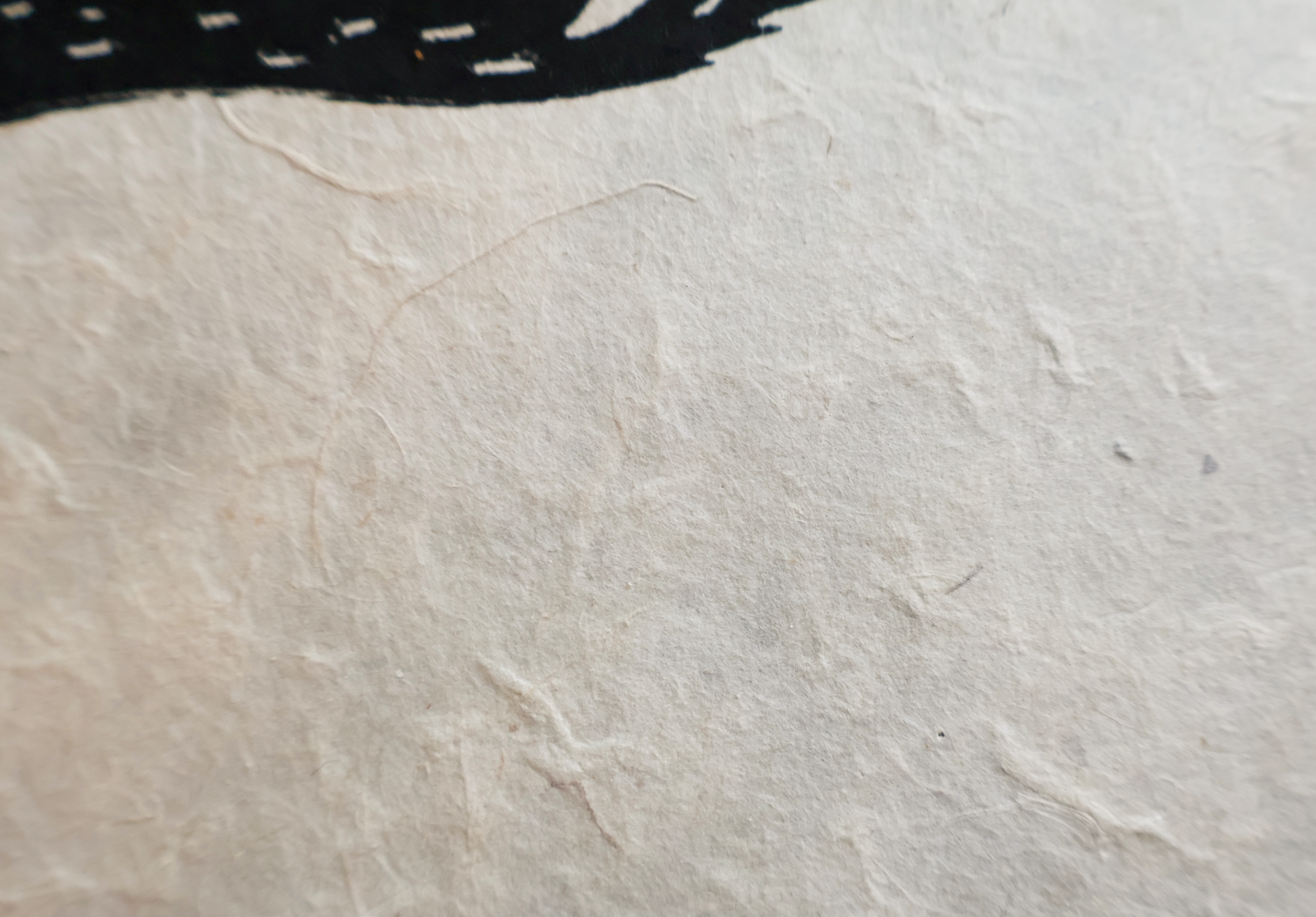 A close-up of textured off-white handmade paper with a dark shadow or object edge visible in the upper left corner.