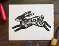 A black and white art print of a leaping rabbit with floral and swirl patterns, displayed on a dark wooden table with red and yellow tools partially visible at the edges.