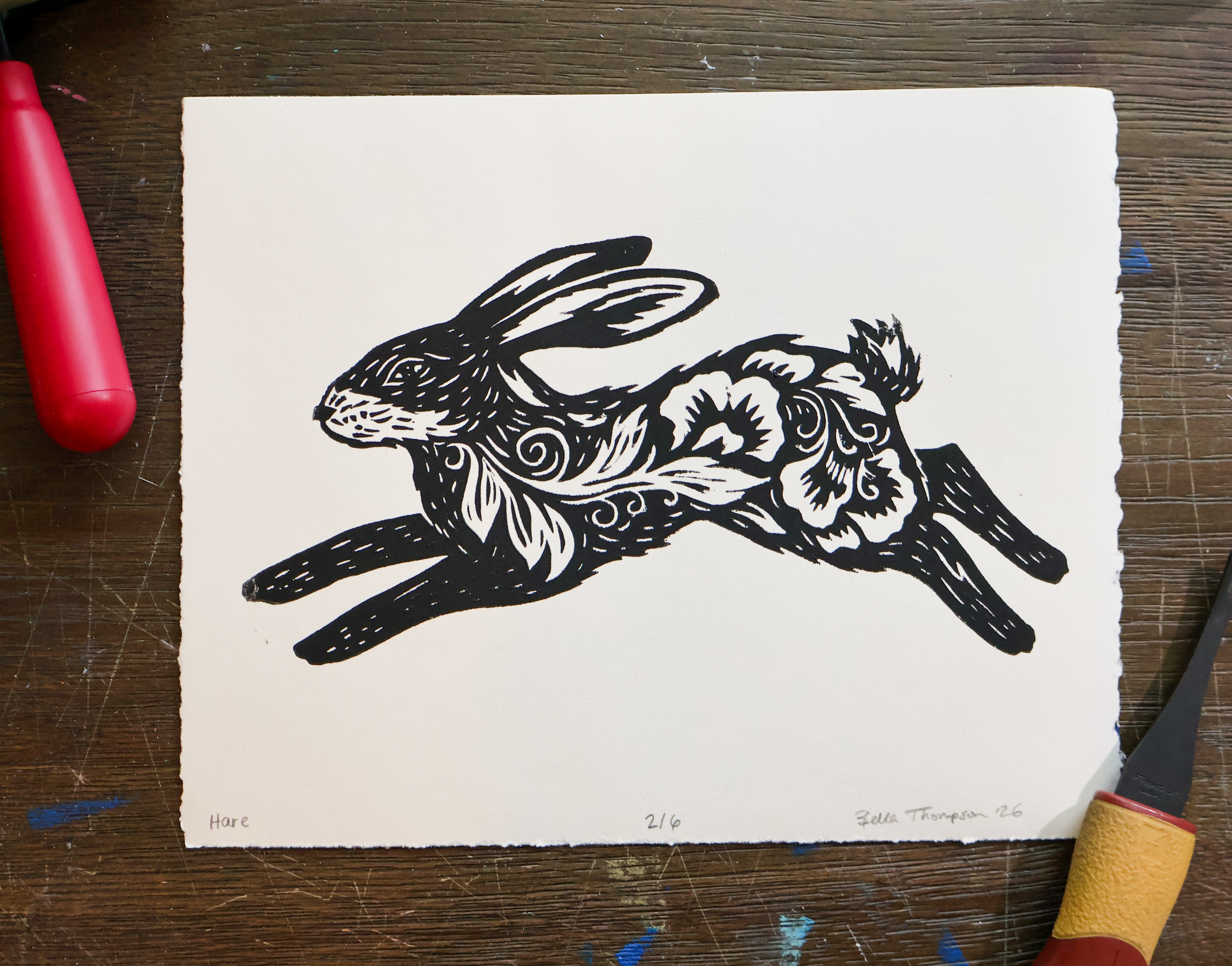A black and white art print of a leaping rabbit with floral and swirl patterns, displayed on a dark wooden table with red and yellow tools partially visible at the edges.