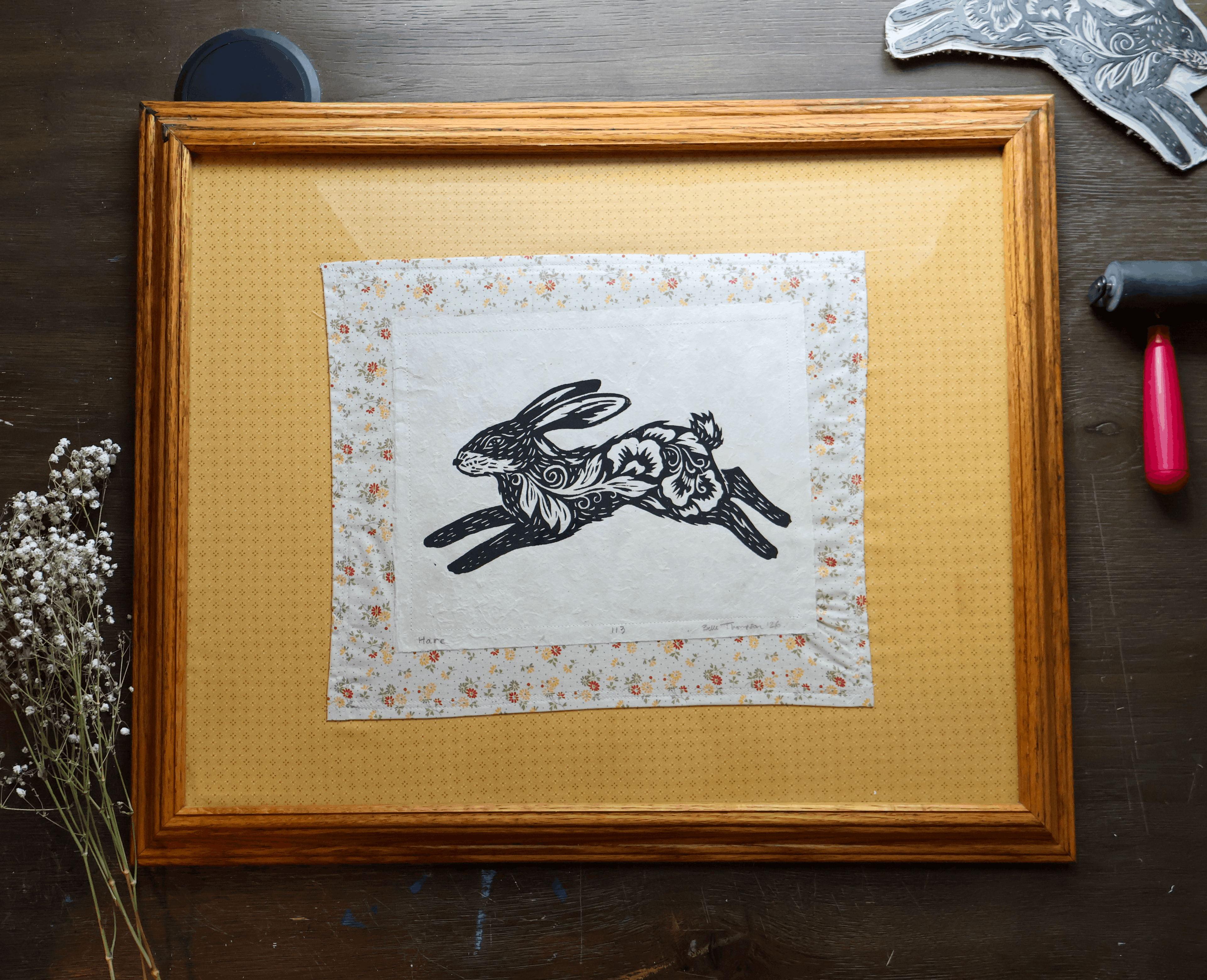 A framed art print featuring a black ink rabbit illustration on a white fabric background with a floral border, set inside a wooden frame on a dark wood surface with decorative props.
