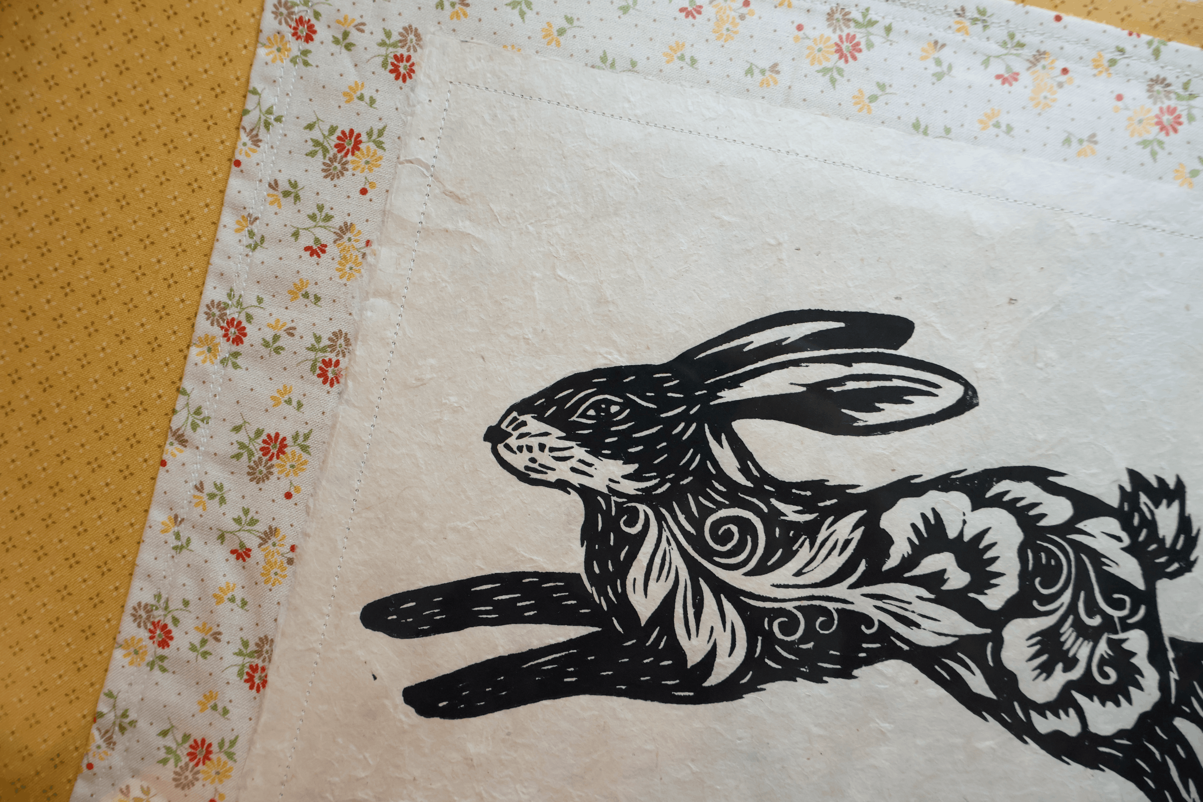 A close-up of a white fabric textile with a black rabbit illustration and a floral border, laid on a yellow textured surface.