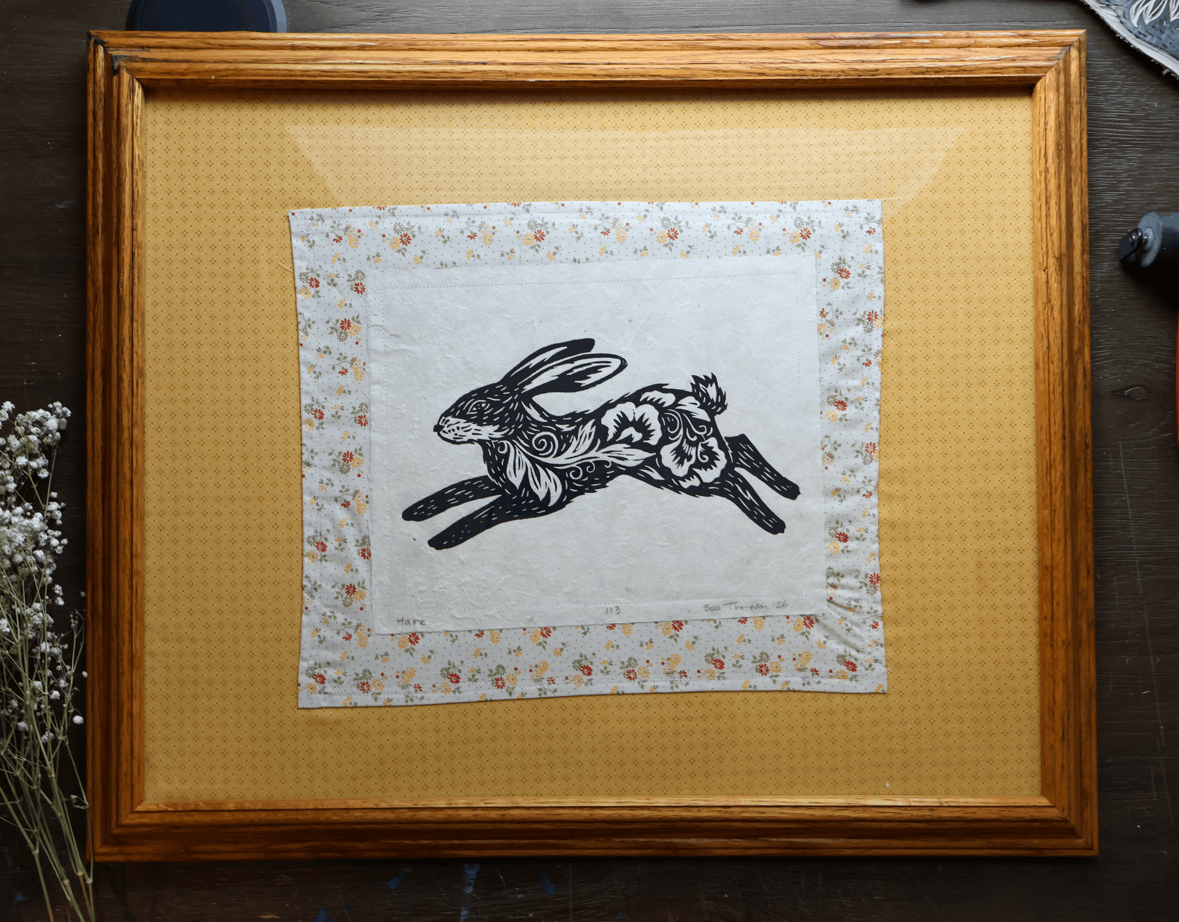 A framed art print featuring a black ink rabbit illustration on a white background with a delicate floral border, set inside a warm brown wooden frame with a yellow mat, displayed on a dark wooden surface.