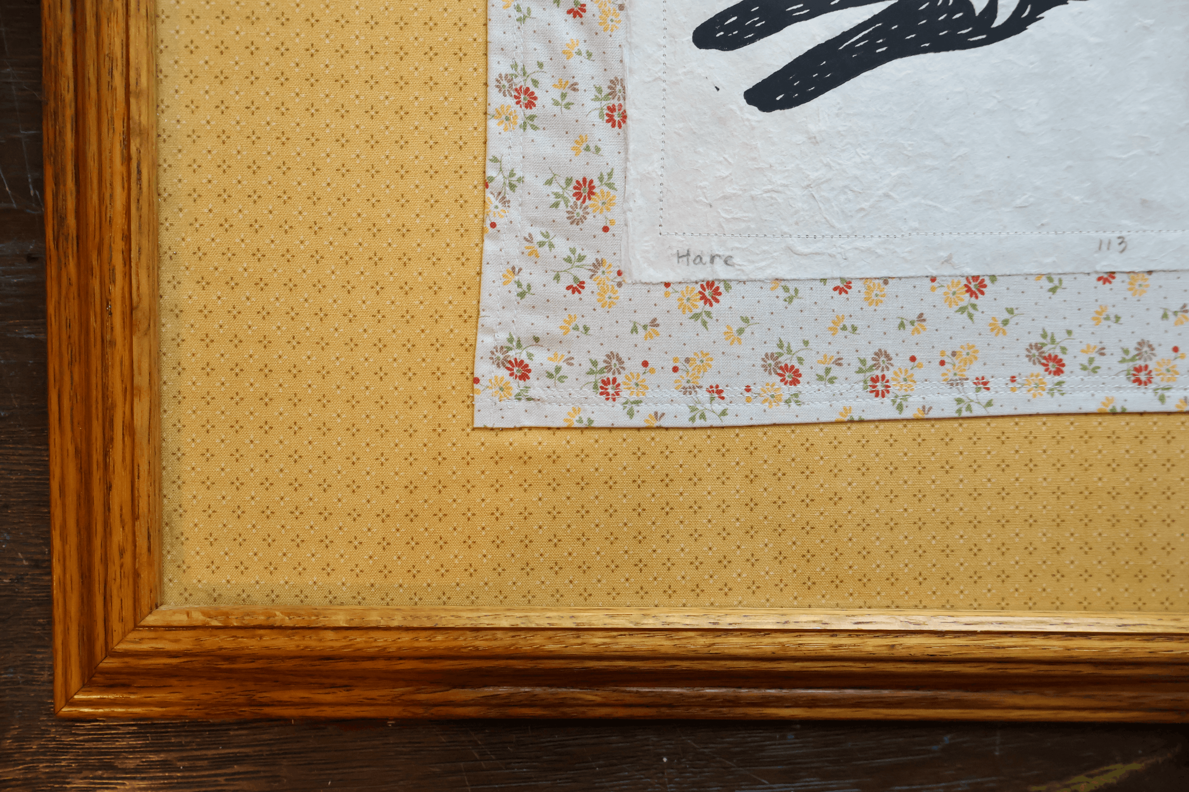 Close-up of a framed textile art piece featuring a floral patterned border, a textured cream center, and a wooden frame on a dark wood surface.