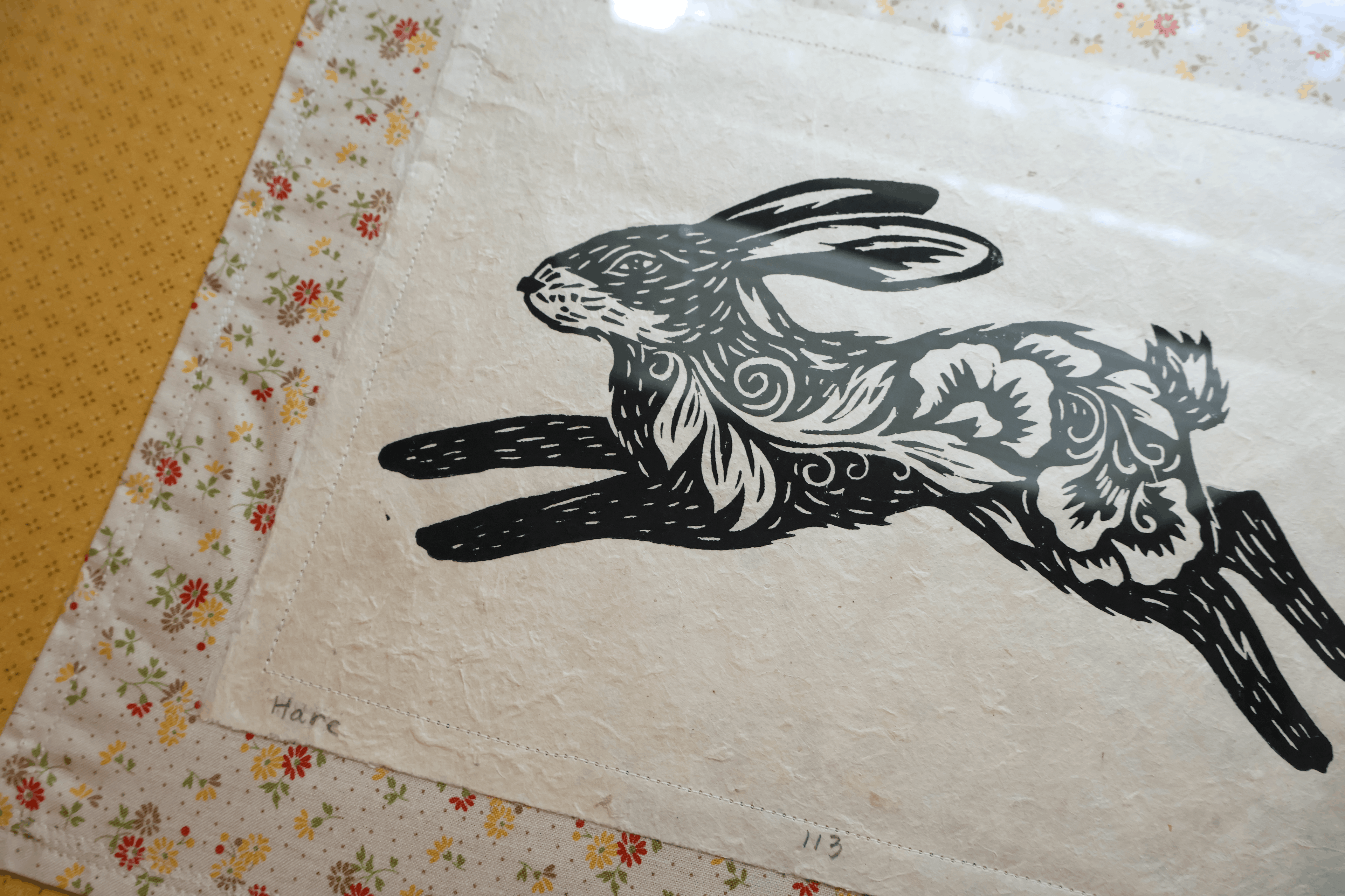 A square handmade paper art print featuring a black ink illustration of a leaping rabbit with ornate floral and swirl patterns. The print is displayed on a floral-patterned fabric border next to a yellow textured surface.
