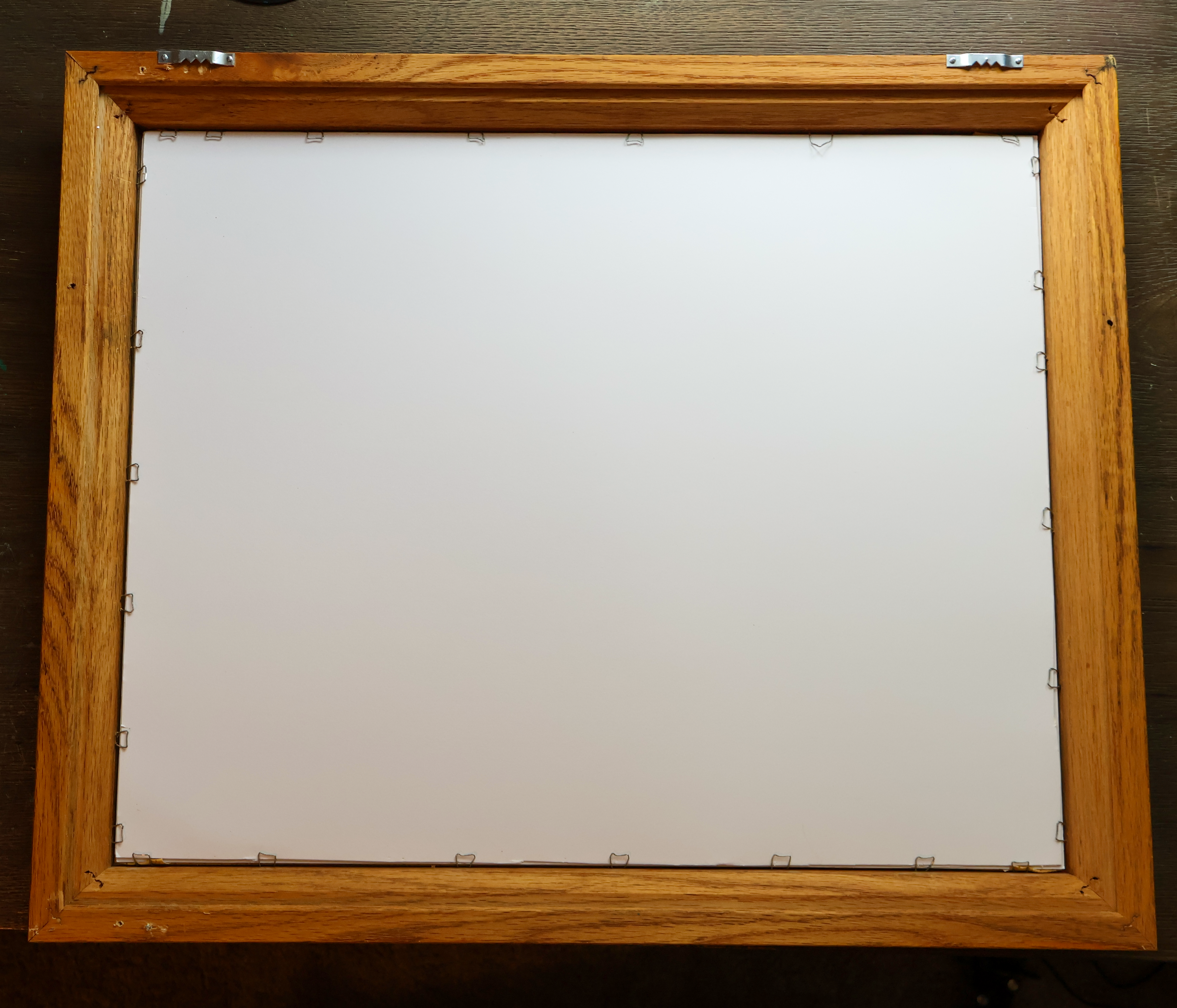 A square wooden picture frame with a natural oak finish, shown from the back with mounting pins visible around a blank white backing board, resting on a dark wooden surface.