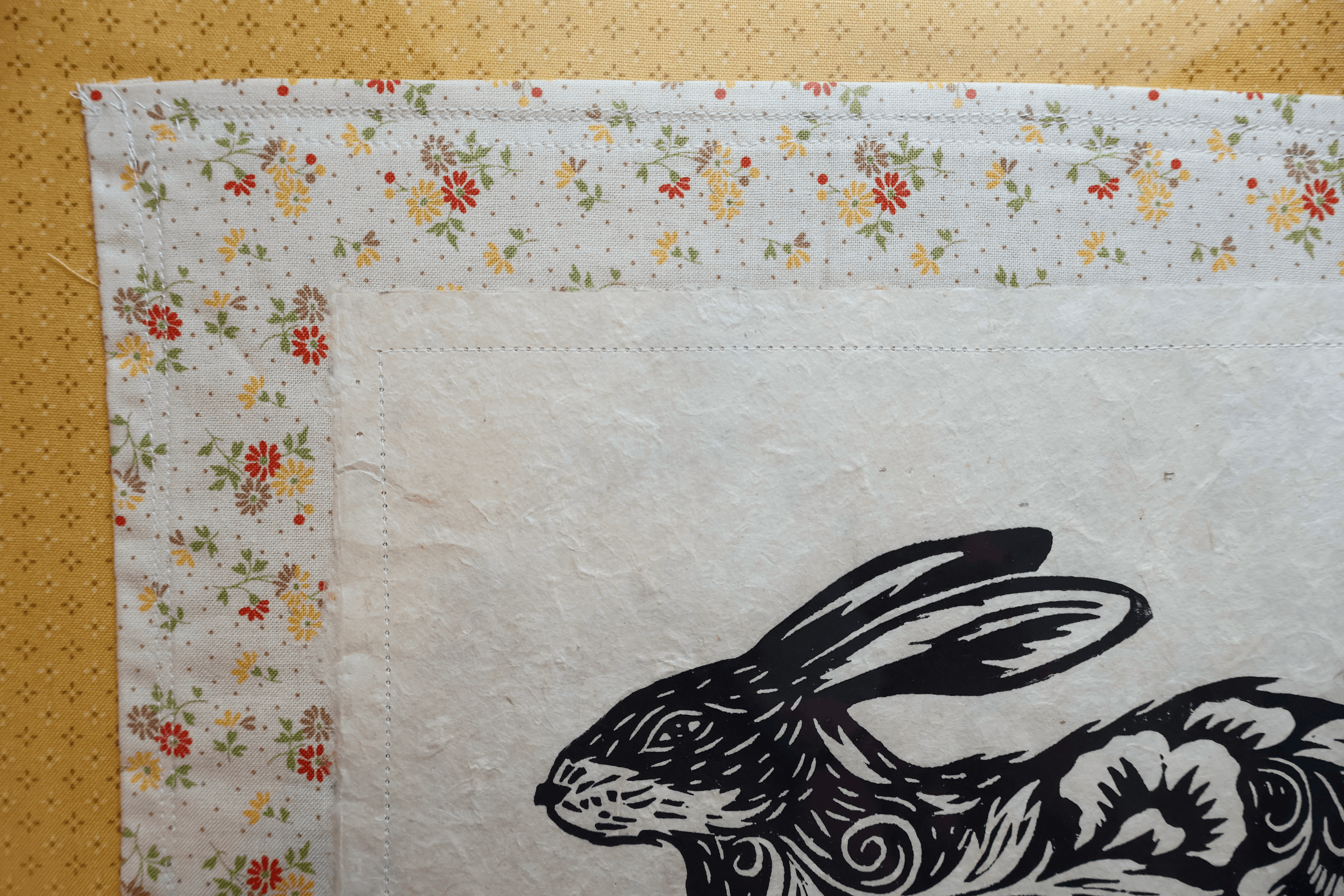 A close-up of a white fabric textile with a floral border and a black rabbit illustration, laid flat on a tan textured surface.