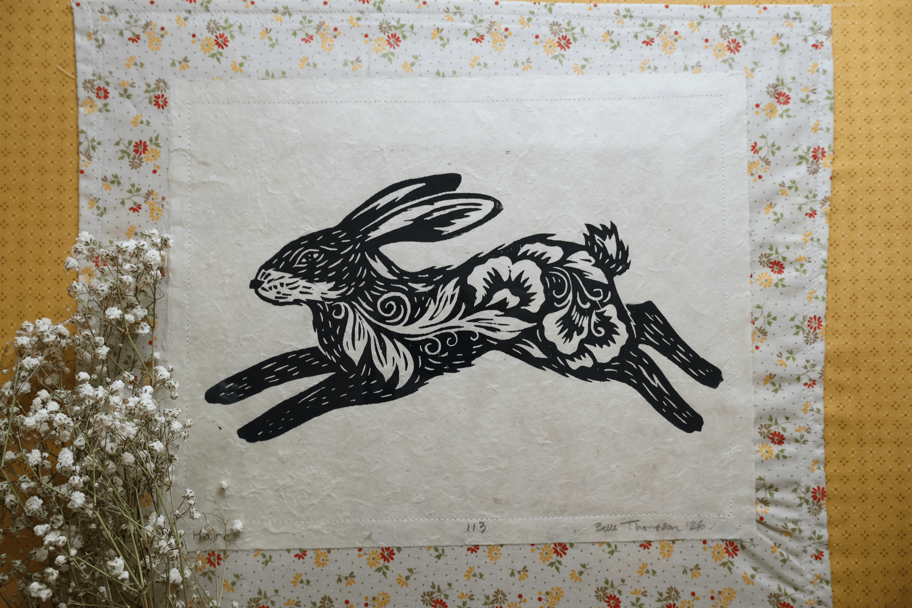 A square textile or art print featuring a black ink rabbit illustration with floral and swirl patterns on an off-white center panel, surrounded by a floral border. The piece is laid flat on a yellow textured surface with white baby's breath flowers at the lower left.