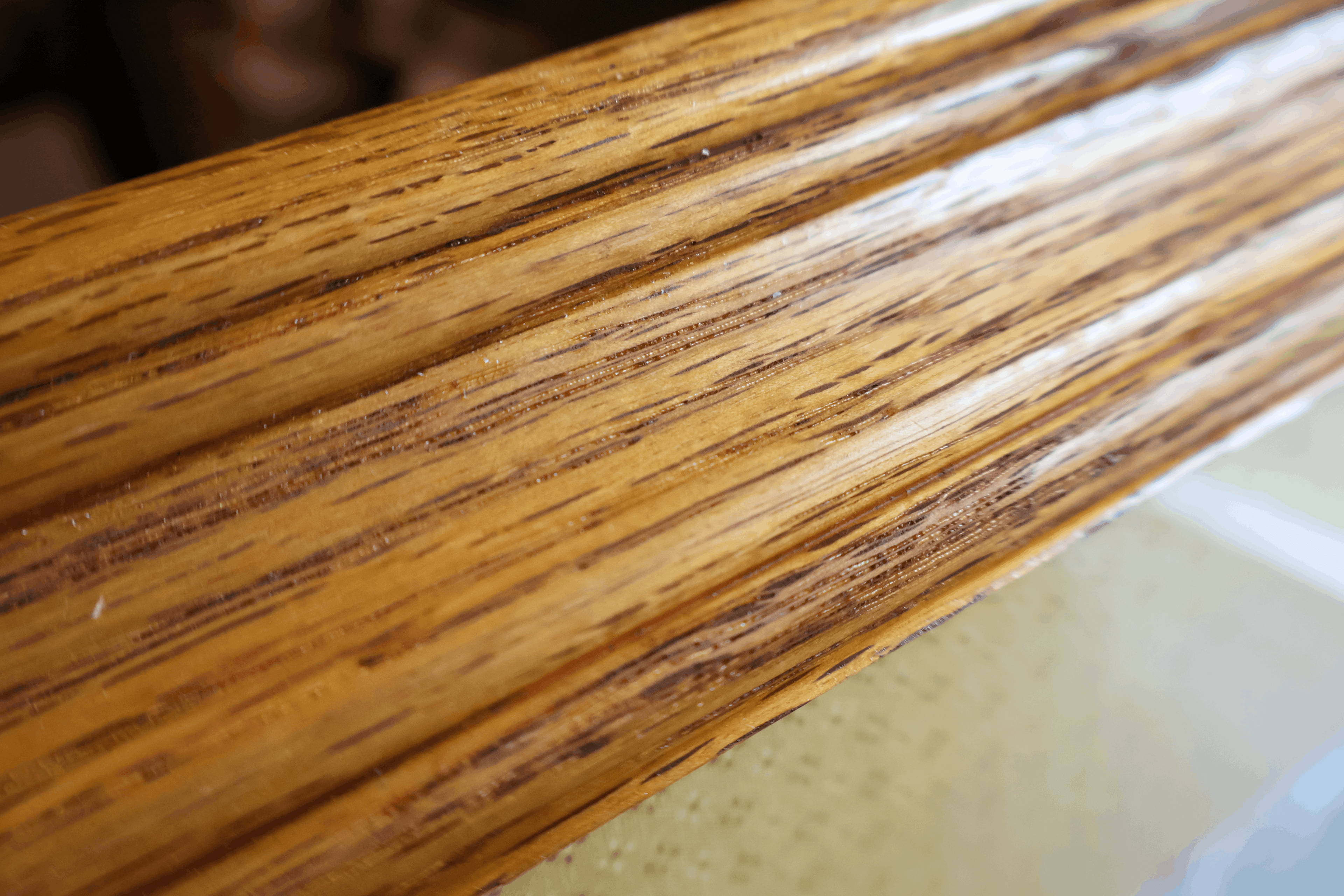 Close-up of a glossy wood grain surface, likely a furniture trim piece or tabletop edge, showing warm brown and tan tones with dark streaks.