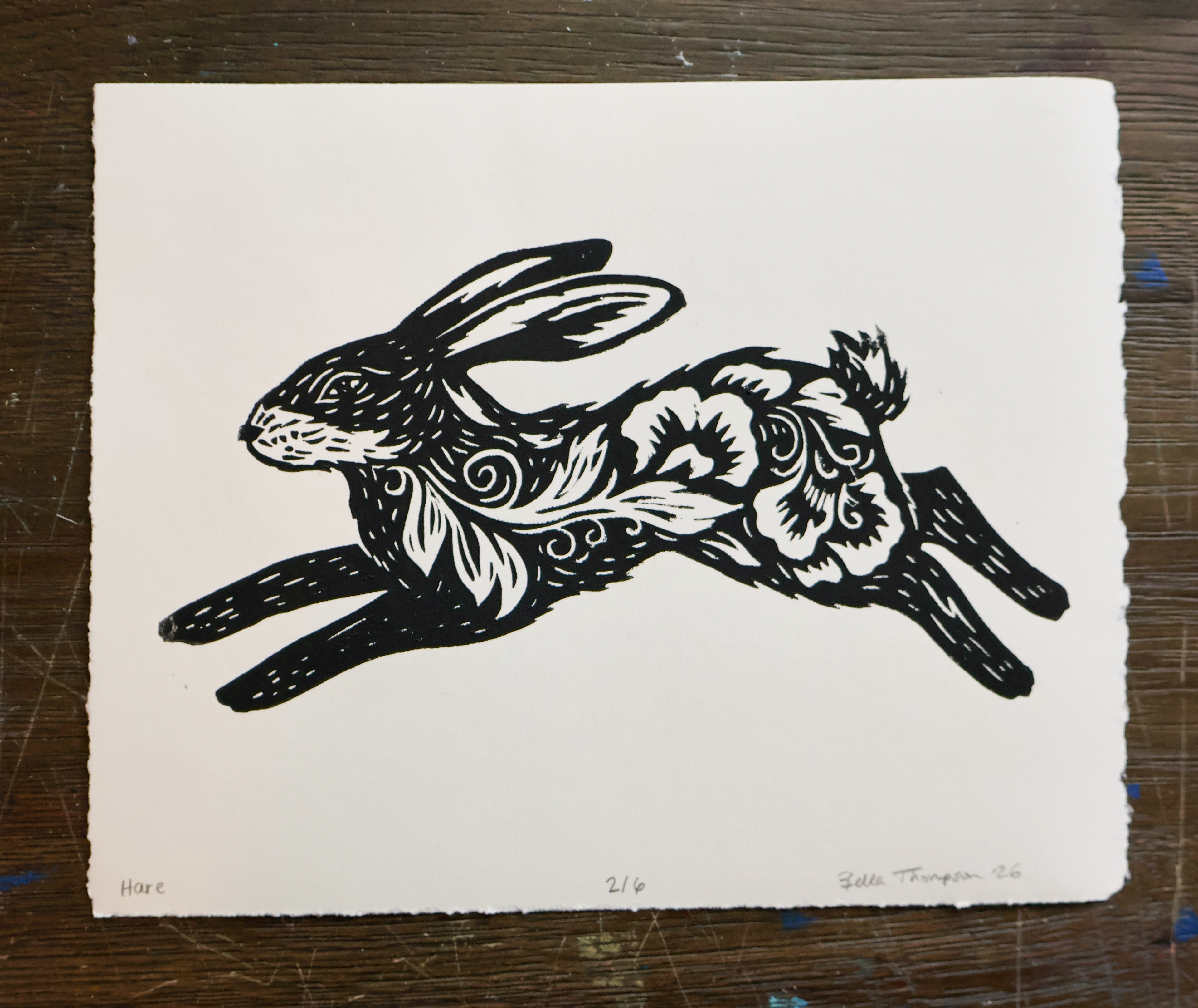 A black ink linocut print of a leaping rabbit on textured off-white paper, displayed flat on a dark wooden surface.