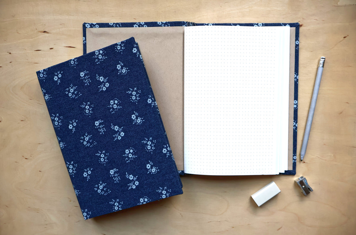 A handmade fabric-bound notebook with a navy blue floral patterned cover, shown both closed and open on a light wood desk with a pencil, eraser, and sharpener.