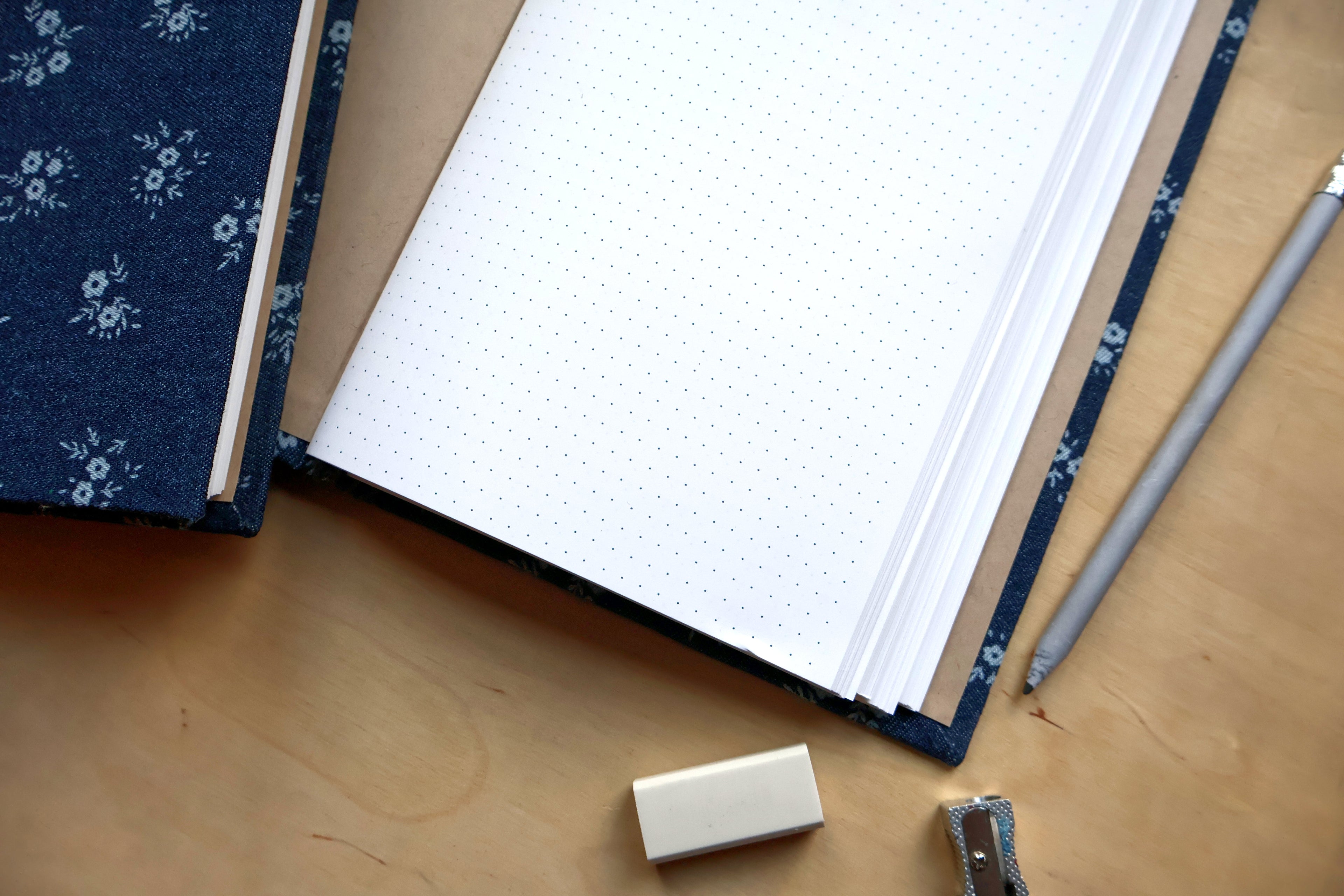 A top-down view of a notebook with white dotted pages and a navy blue floral cover, placed on a wooden desk with a pencil, eraser, and other stationery items.