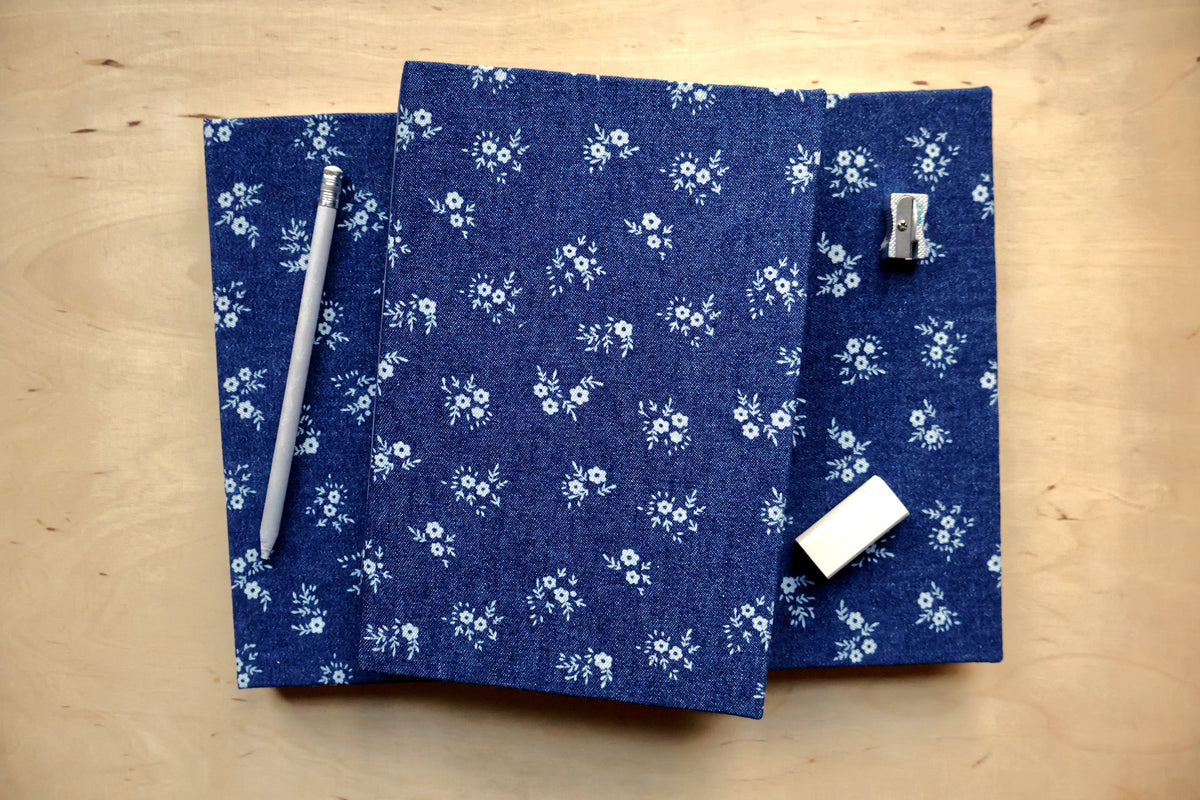 Two blue fabric-covered notebooks with a white floral pattern are arranged on a light wood background. One notebook has a silver pencil on top, and the other has a silver binder clip and a white eraser.