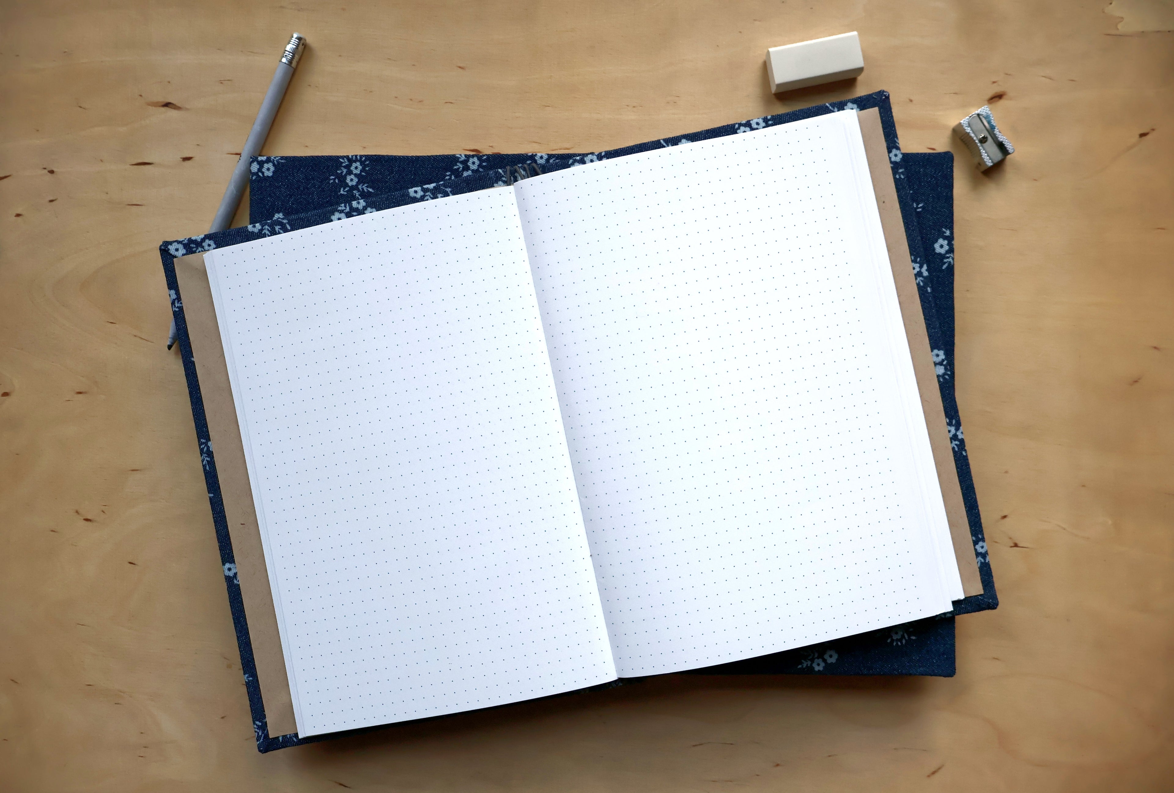An open notebook with white graph paper pages lies on a wooden desk, surrounded by a pencil, eraser, and small paper clips. The notebook has a dark navy blue floral-patterned cover.