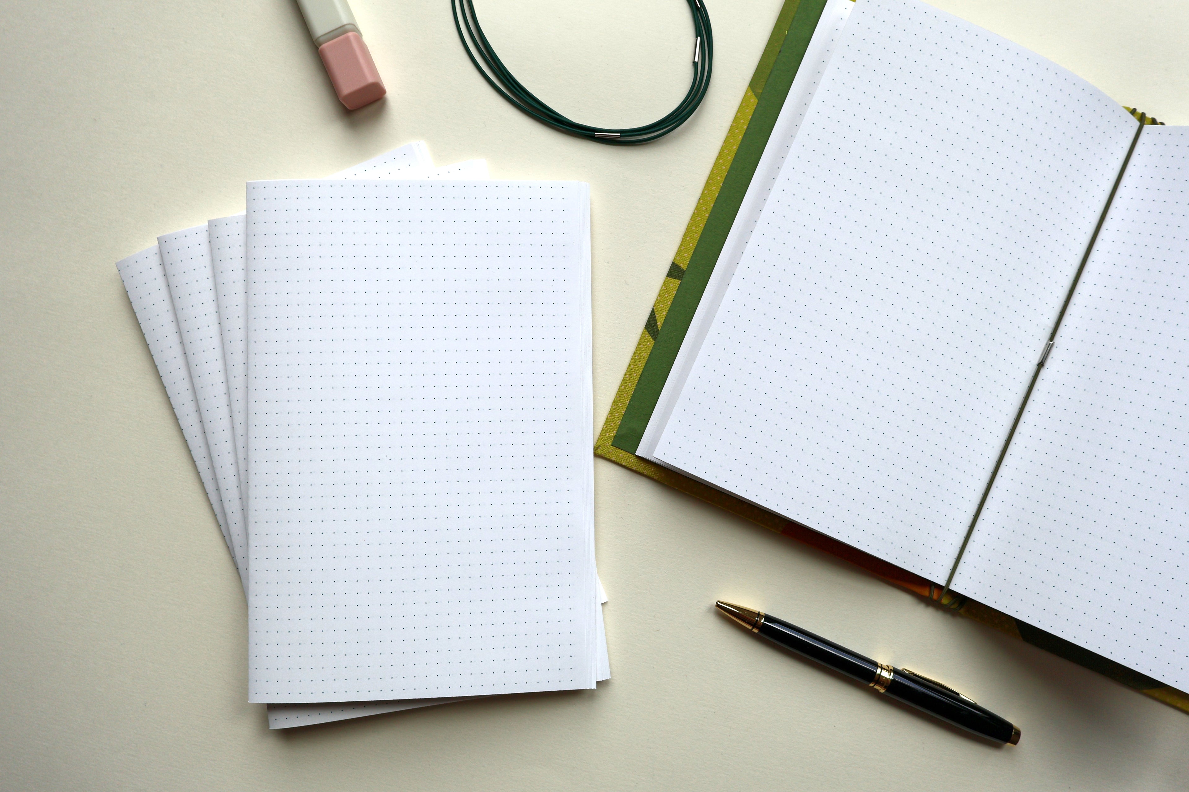 A styled flat-lay of stationery items on a light beige surface: an open navy-bound bullet paper journal with a black and gold pen, several loose bullet paper sheets, a pink-and-white highlighter, and a dark green loop of elastic journal binders. Showcasing Dovetail Studio's handmade refillable journal refills
