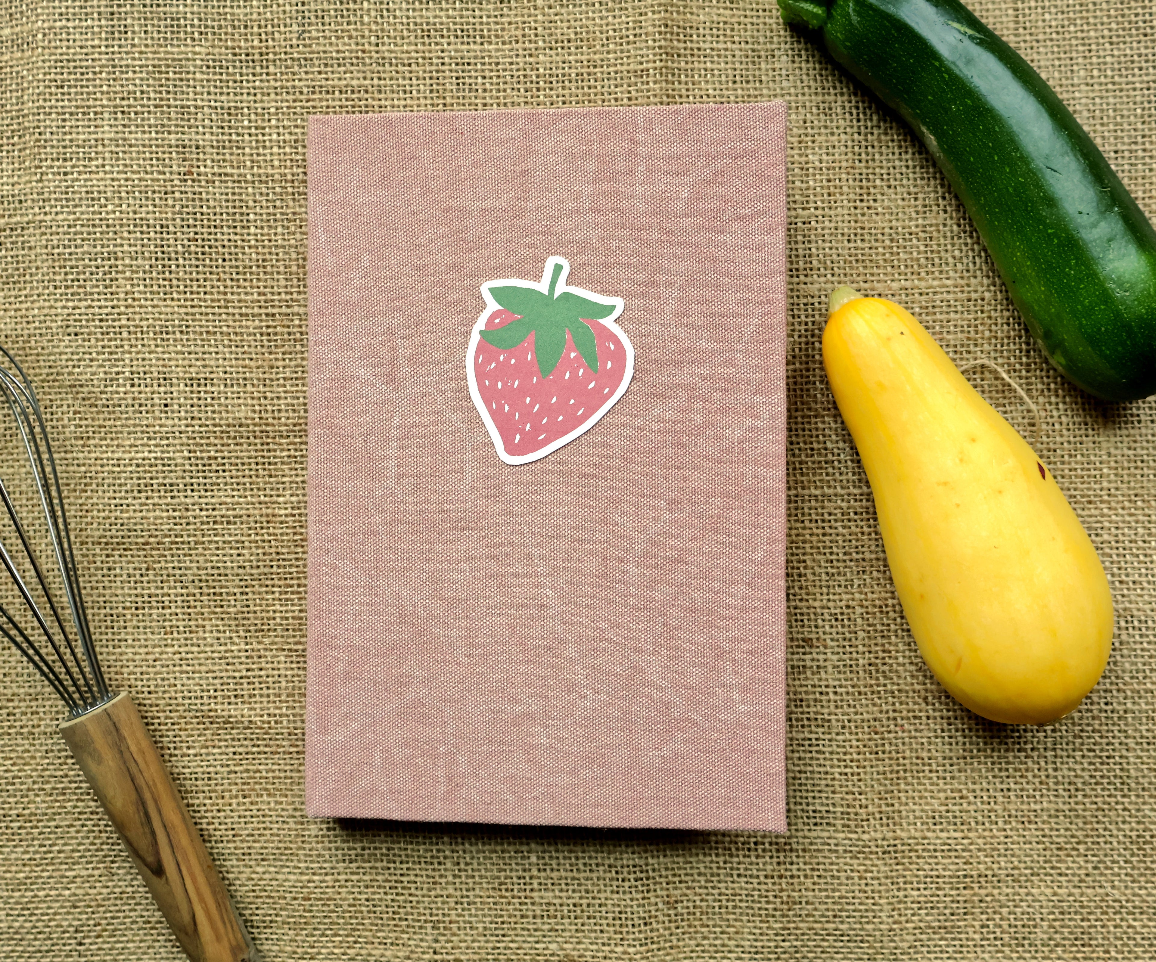 A pink textured hardcover recipe log with a strawberry linocut print design on the cover, shown next to an open matching book on a burlap background with a green cucumber and a yellow squash as props.