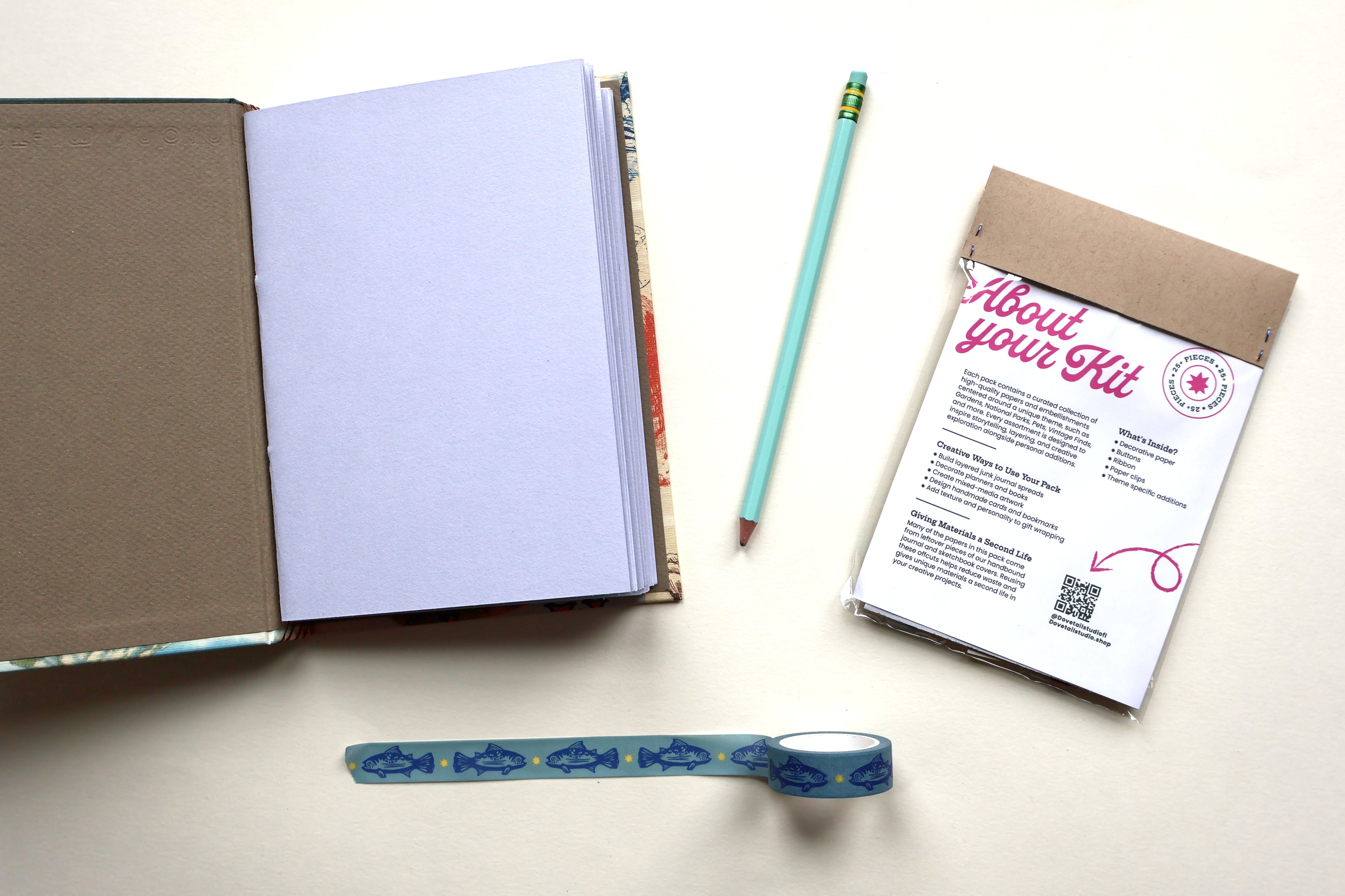 A flat-lay view of a DIY junk journaling kit on a light beige background, including an open sketchbook with blank pages, a mint green pencil, a roll of blue trout patterned washi tape, and a small junk journaling scraps kit labeled 'About your Kit'.