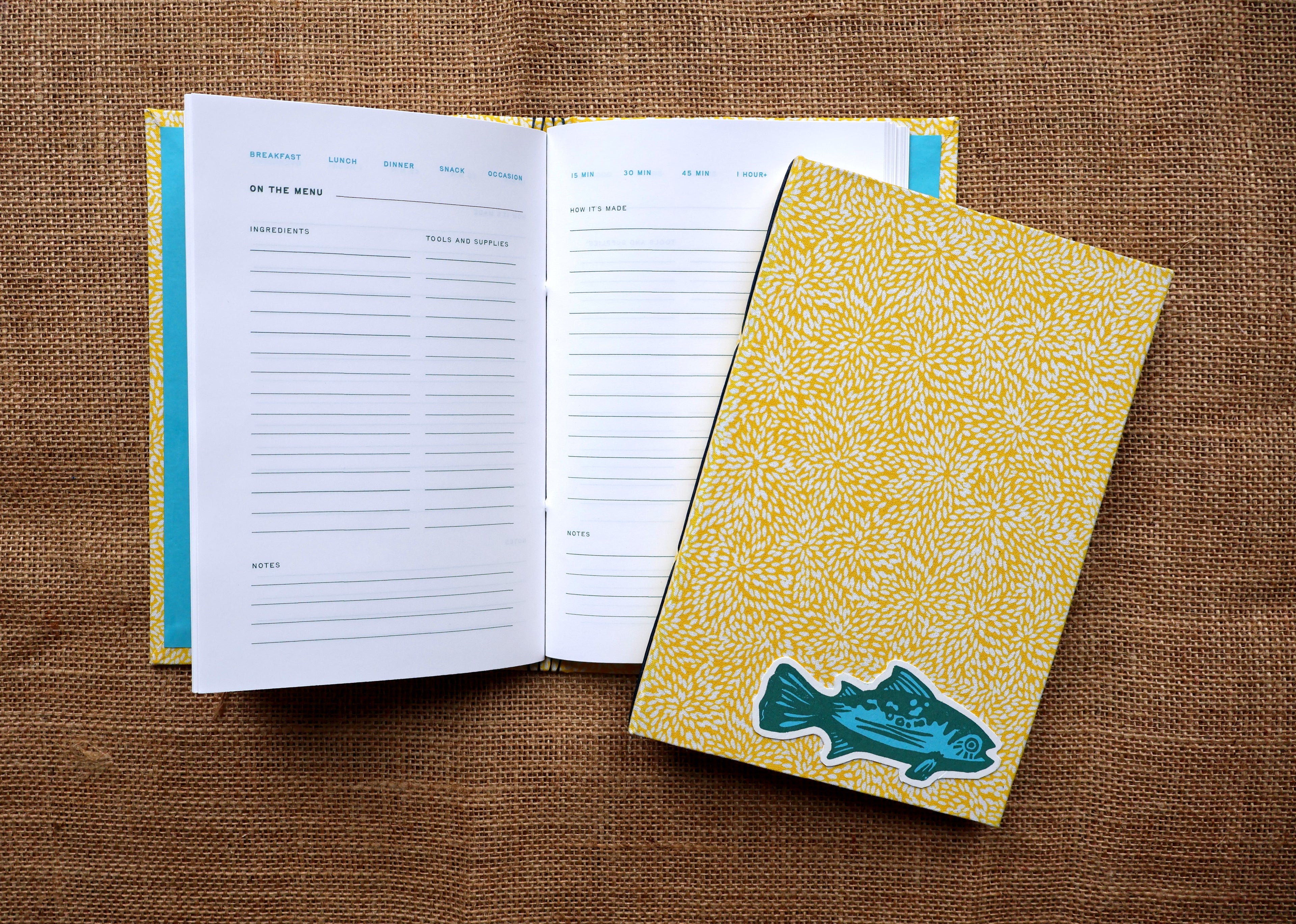 A yellow patterned recipe log cover with a teal fish linocut design, shown next to an open matching recipe log on a brown burlap background.