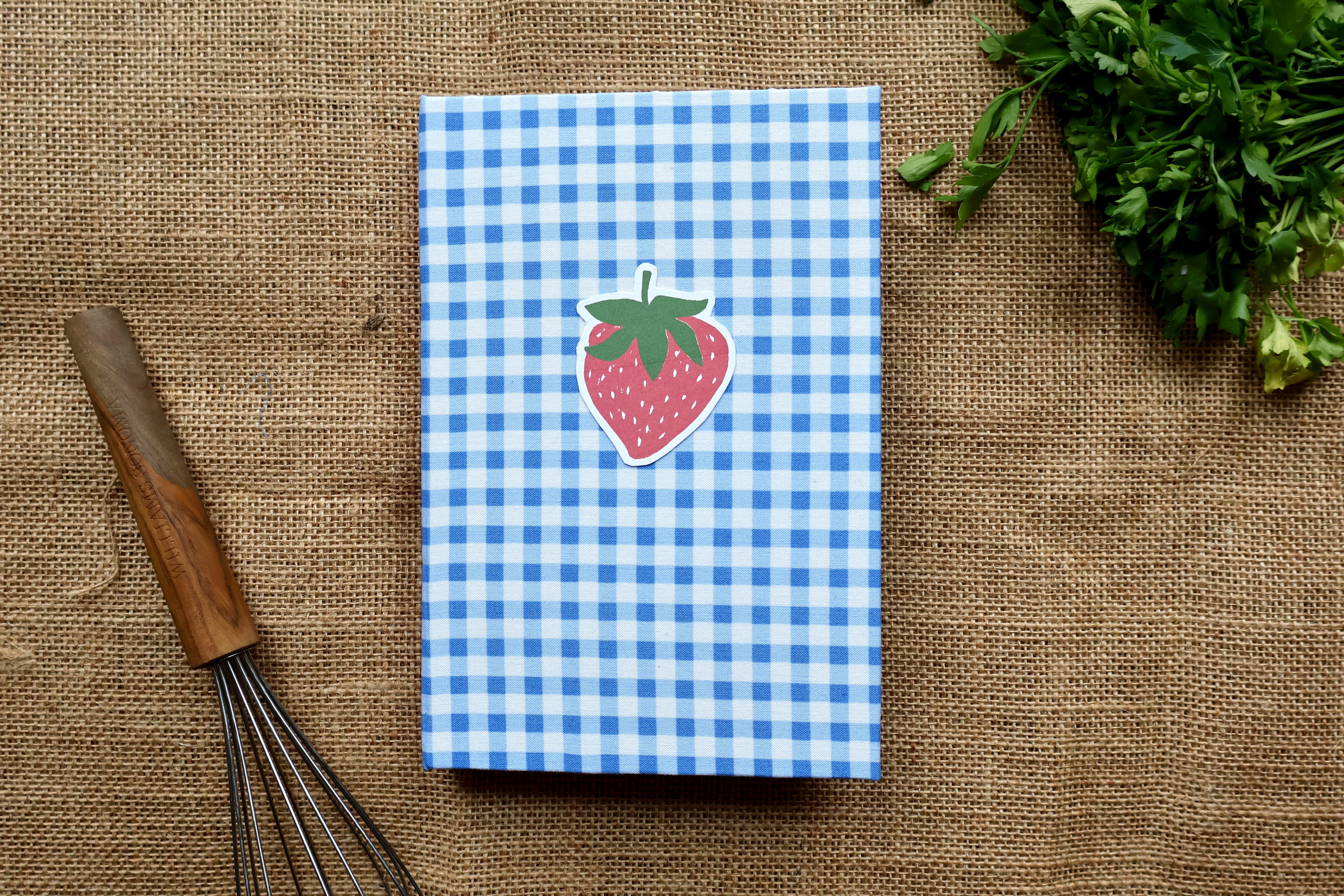 A recipe log with a blue and white gingham pattern cover and a strawberry linocut design on the front, displayed on a burlap surface with a wooden whisk and parsley.