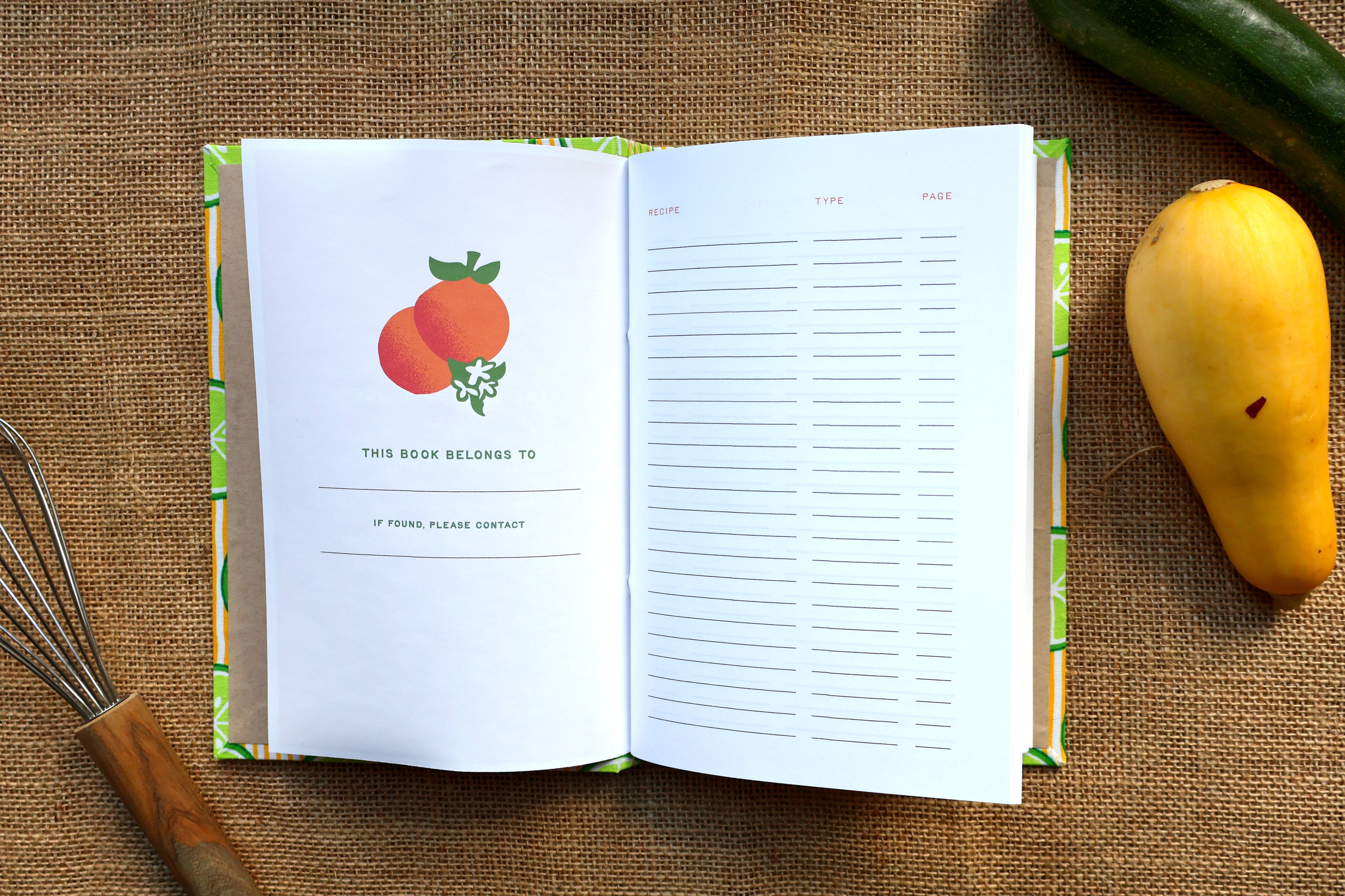 An open hardcover recipe log lies on a burlap surface, with the left page showing a fruit illustration and the right page having lined writing space. A whisk is at lower left, a dark green cucumber is at upper right, and a yellow squash is at right.