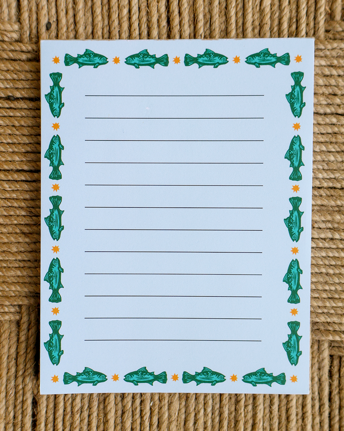A lined writing pad with a fish-themed border, placed on a woven straw surface. The pad has horizontal lines in the center and green fish illustrations with small orange stars around the edges.