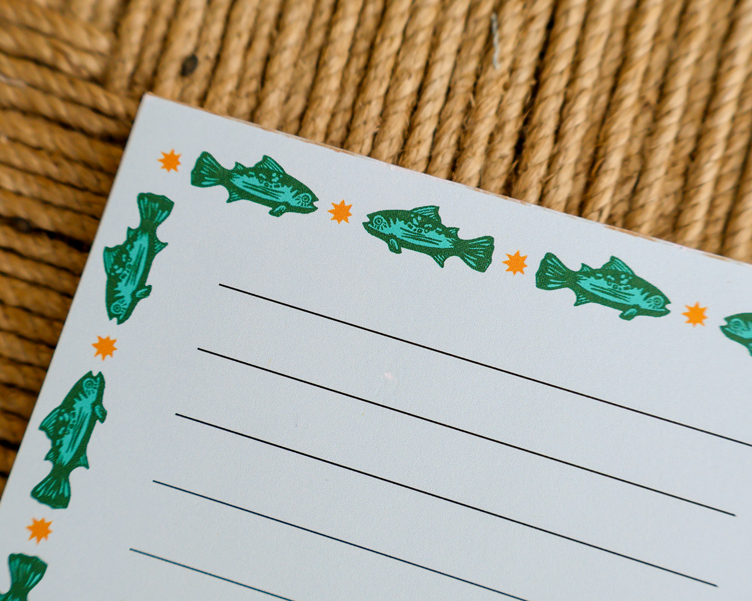 A close-up of a white lined writing pad or notepad with green fish illustrations and small orange star accents along the border, placed on a woven tan surface.