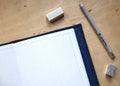 An open graph notebook with a dark navy blue cover, placed on a light wood desk alongside a gray mechanical pencil, a white eraser, and a silver sharpening stone.