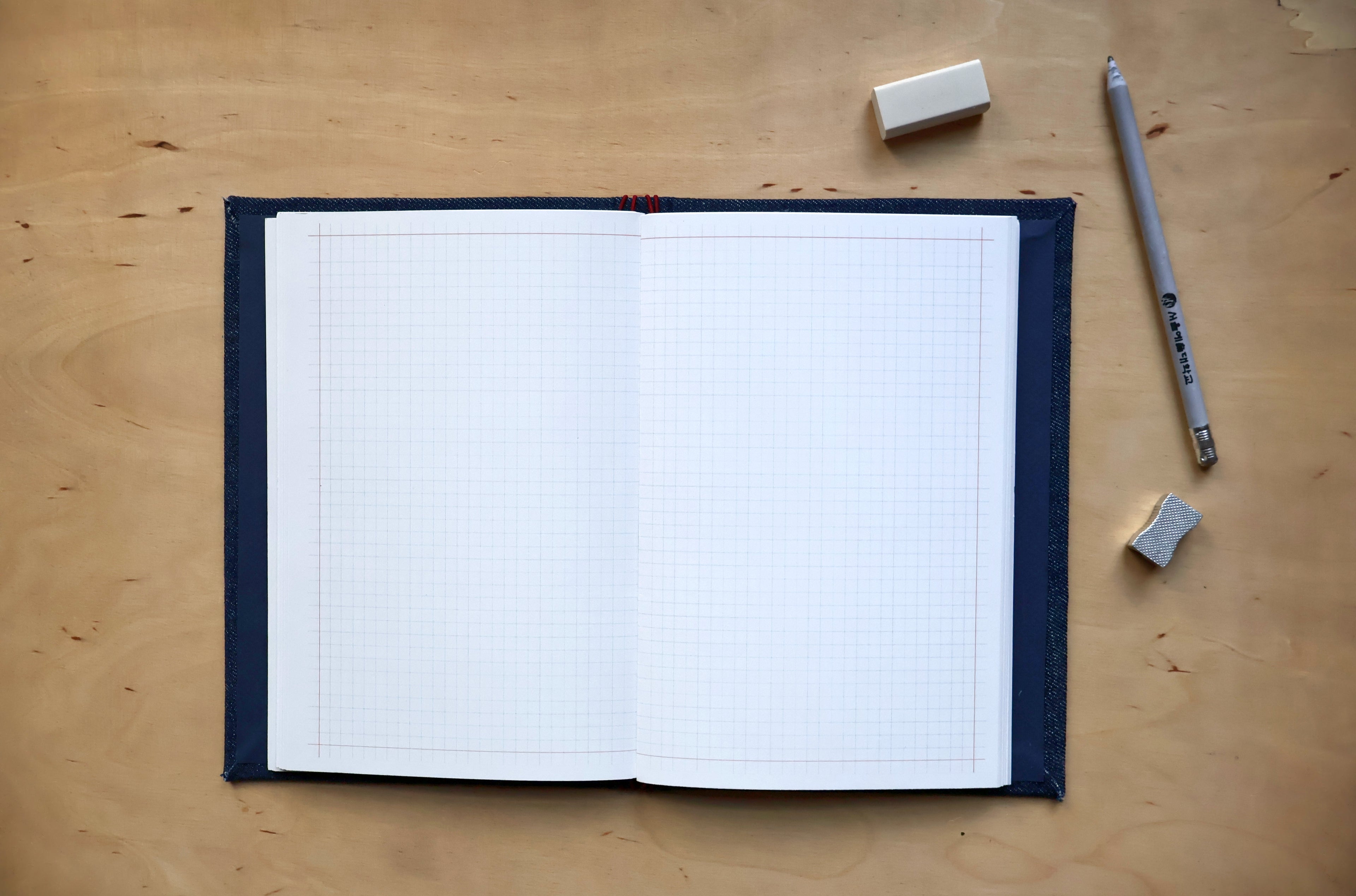 An open graph notebook with white grid paper and a dark navy blue cover, placed on a light wood desk with a pencil, eraser, and small rubber accessory nearby.