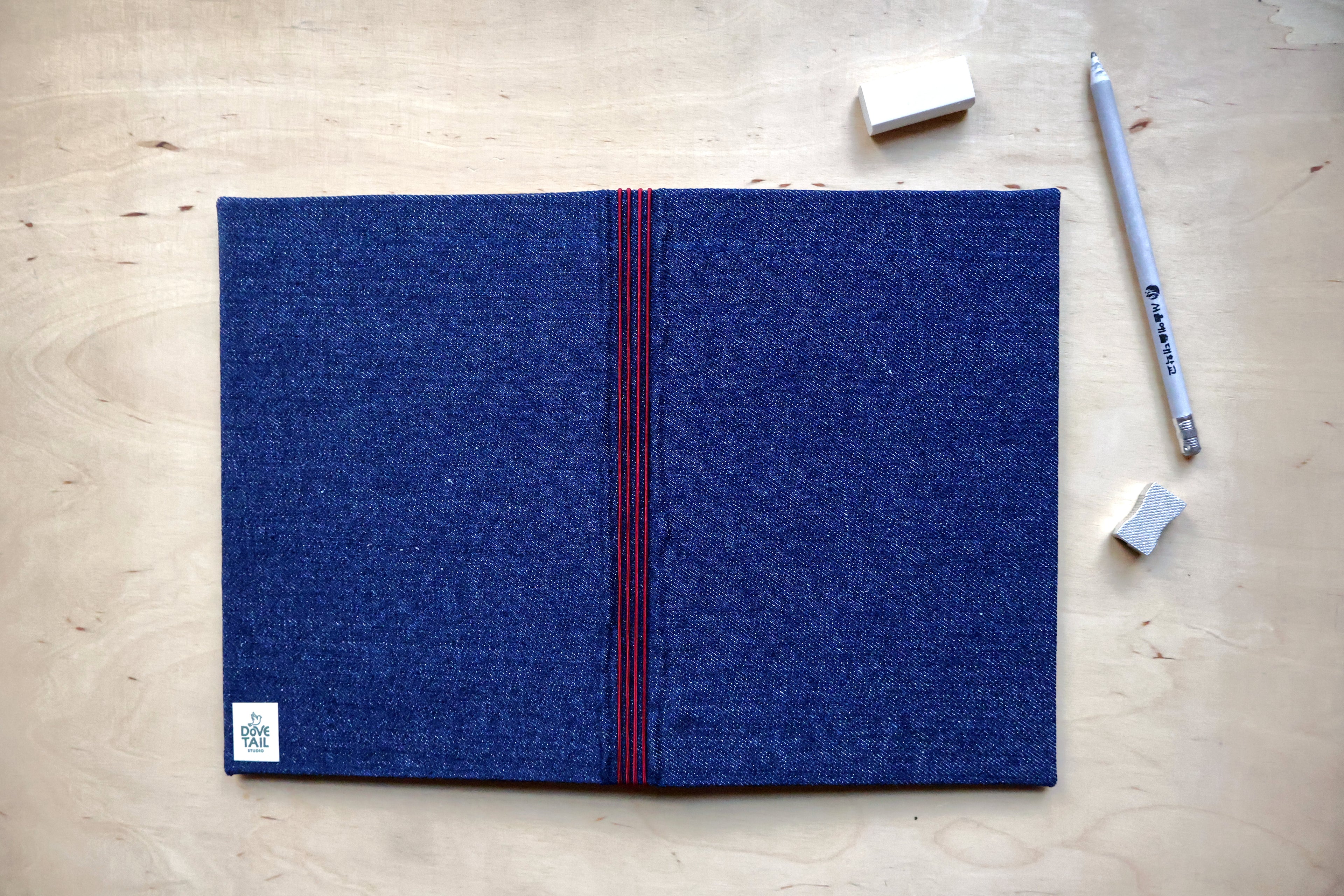 A dark blue fabric journal with a textured weave, featuring a vertical red stripe down the center and a small white logo patch in the bottom-left corner. The notebook is laid flat on a light wood background, surrounded by a silver pen, two small erasers, and a pencil.