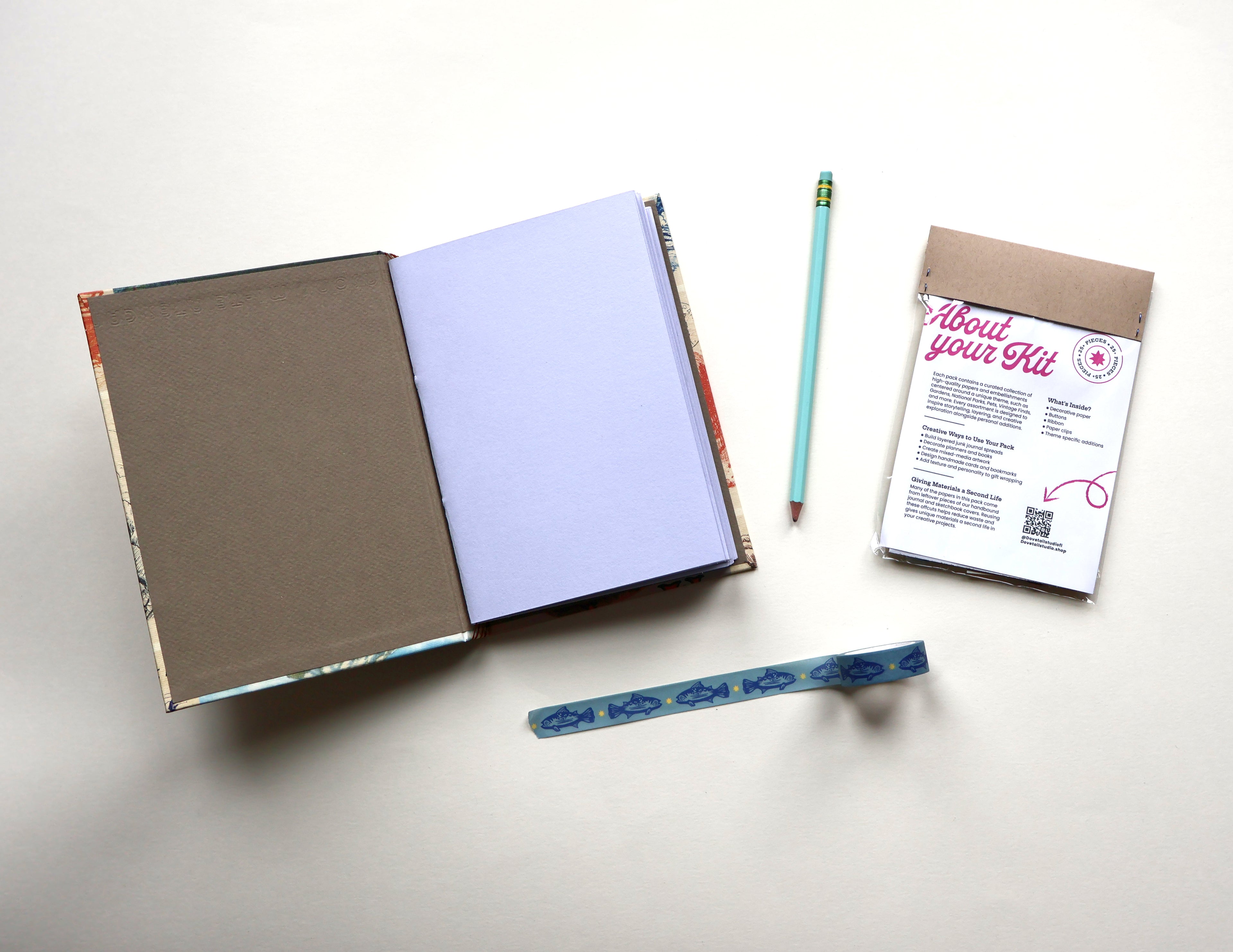 An open sketchbook with a brown cover and blank interior pages, shown on a light neutral background with a teal pencil, a small kraft envelope, and a printed insert card labeled 'About your Kit.' A blue patterned trout washi tape lies below the items.