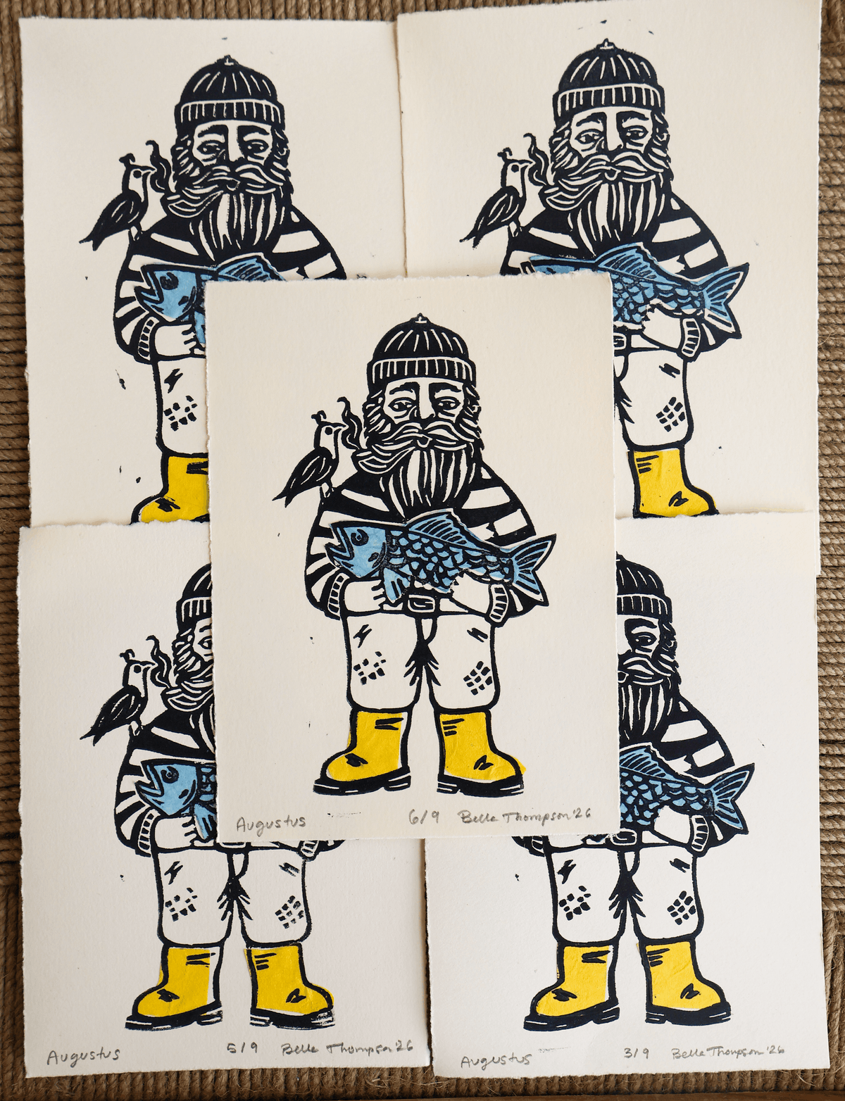 Five art prints featuring a stylized bearded figure in winter clothing holding a blue fish, arranged on a brown textured surface. The prints are mostly off-white with black, blue, and yellow illustrations.