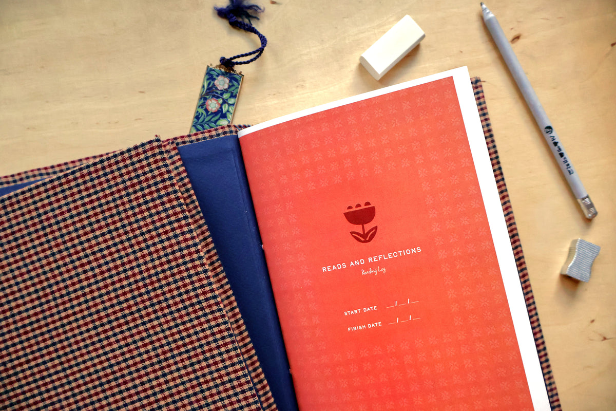 An open reading log on a light wood desk, showing a coral-red interior cover with a tulip logo and the text 'READS AND REFLECTIONS reading log,' alongside a plaid cover, blue inner lining, erasers, and a pencil.