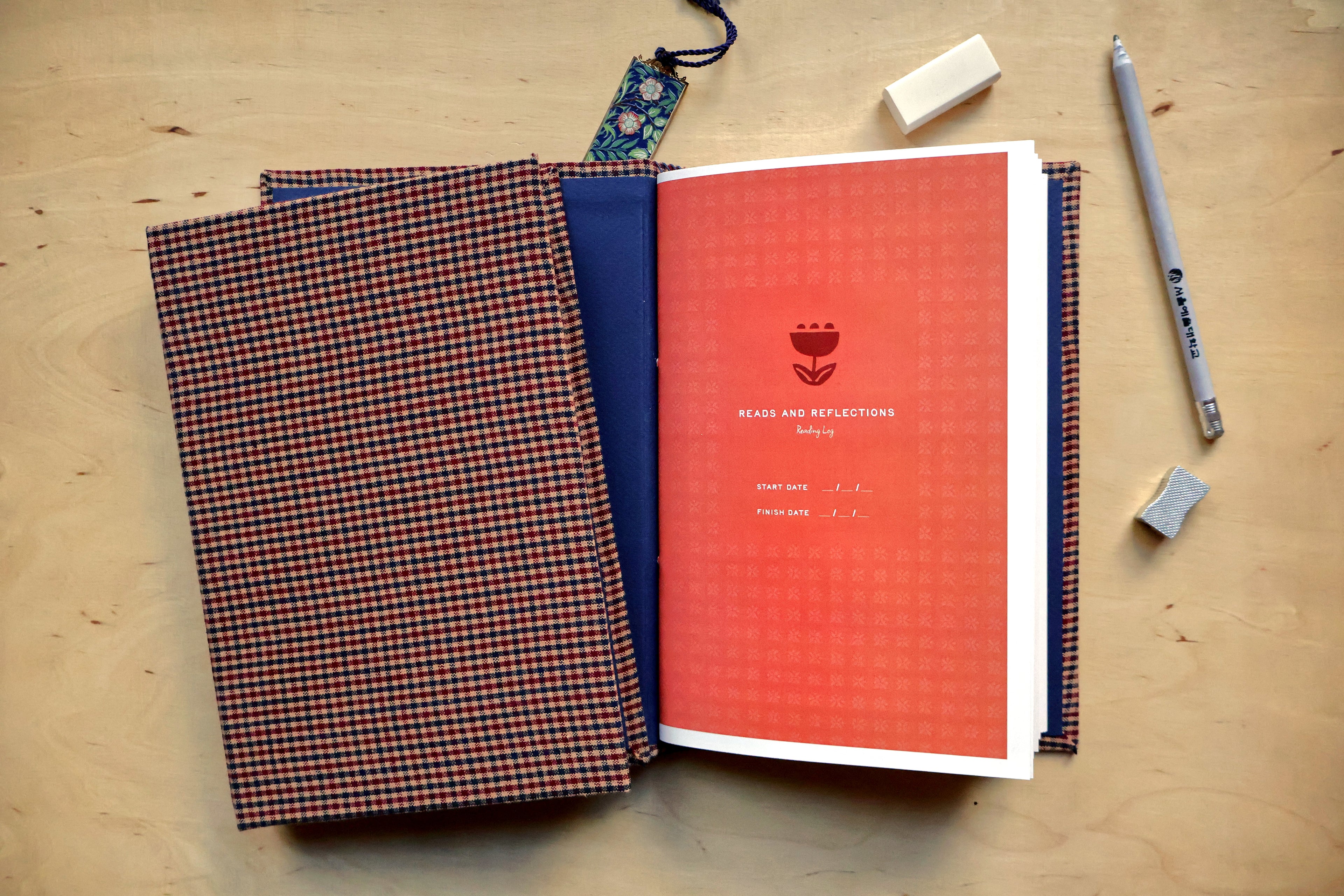 A handmade fabric-bound reading log lies open on a light wood tabletop. The left page has a red, navy, and black checkered patterned cover, while the right page is coral red with a small floral logo and the text 'BEACH AND REFLECTIONS'.