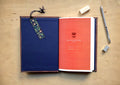 An open reading log laid flat on a light wood surface, with a dark blue cover on the left, a coral-red interior page on the right, a floral bookmark hanging from the cover, and a pen and erasers nearby.