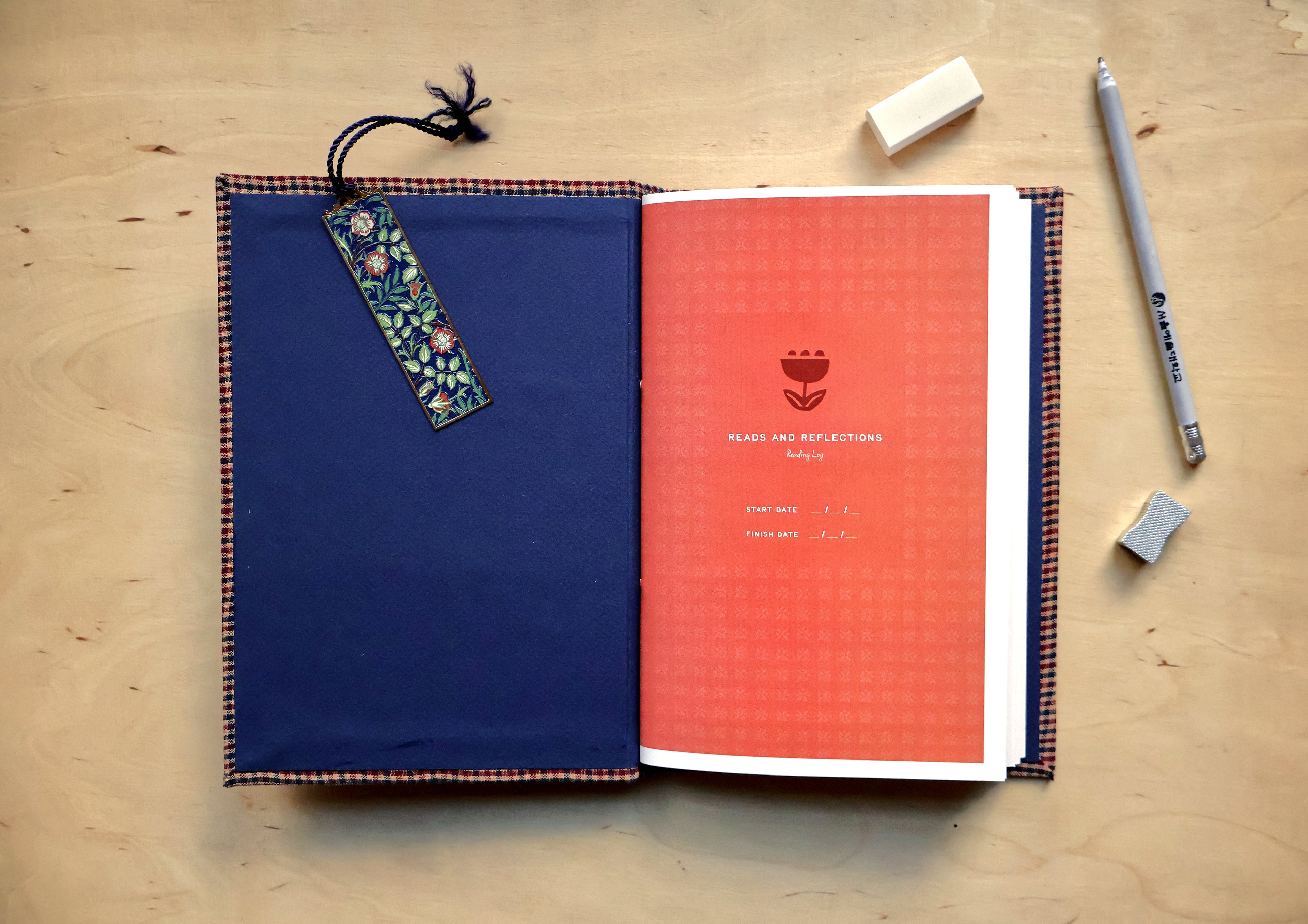 An open reading log laid flat on a light wood surface, with a dark blue cover on the left, a coral-red interior page on the right, a floral bookmark hanging from the cover, and a pen and erasers nearby.