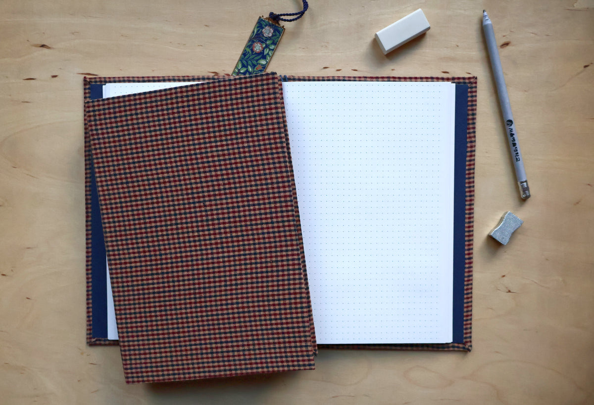 An open bullet journal with a white dotted page, featuring a dark navy blue cover and a brown and orange plaid binding, displayed on a light wood desk alongside a gray pen, an eraser, and a small tag.