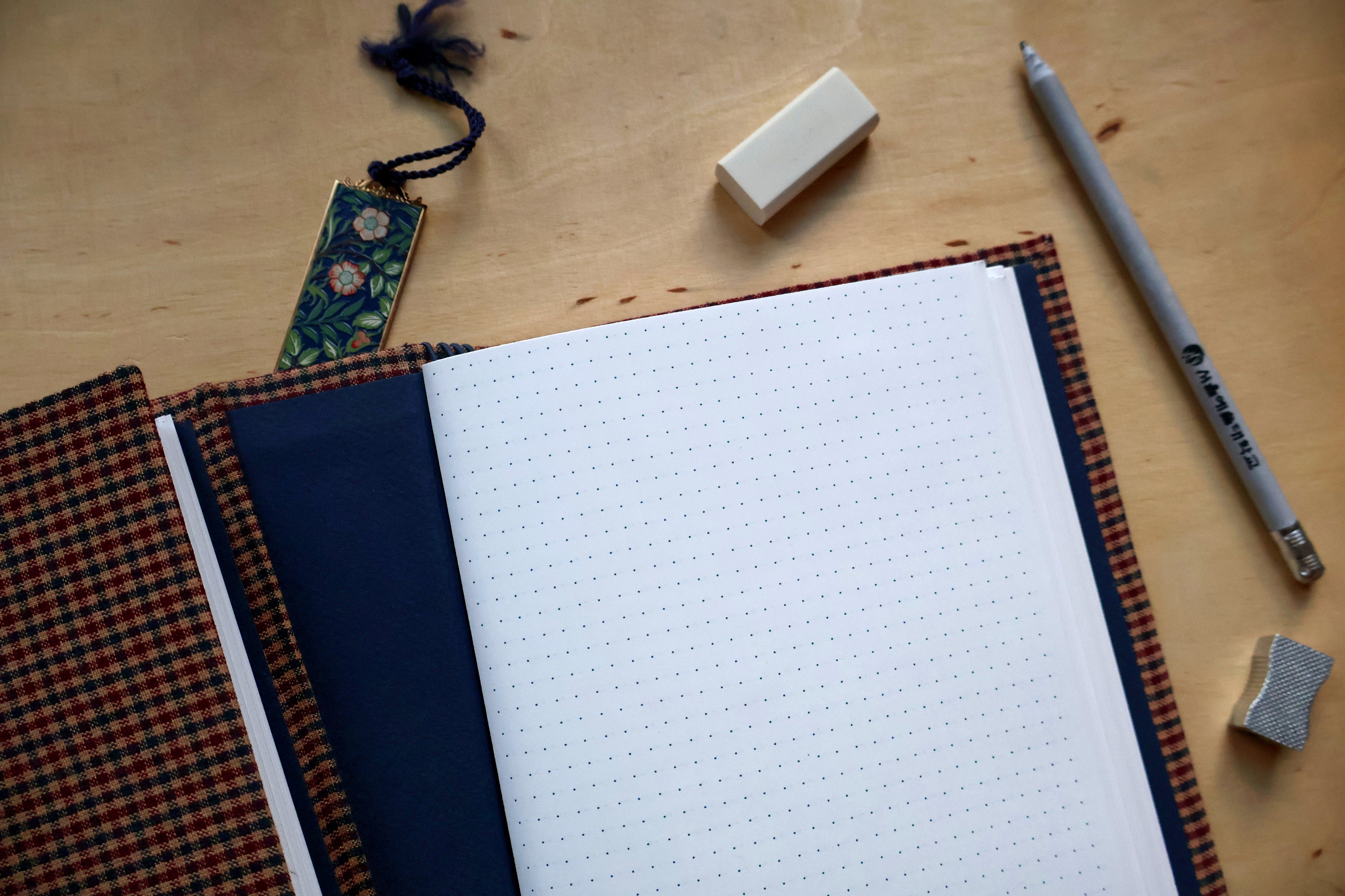 An open bullet journal with a white dotted page, featuring a dark navy blue cover and a brown and orange plaid binding, displayed on a light wood desk alongside a gray pen, an eraser, and a small tag.