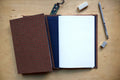 A handmade notebook with a red and black checkered cover, shown open on a light wood desk. The notebook has a dark navy blue inner cover and white graph paper pages.