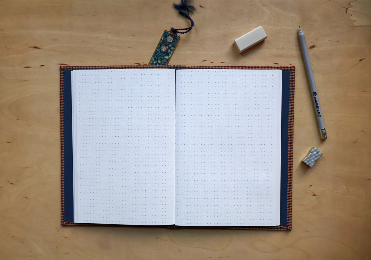 An open bullet journal with a white dotted page, featuring a dark navy blue cover and a brown and orange plaid binding, displayed on a light wood desk alongside a gray pen, an eraser, and a small tag.