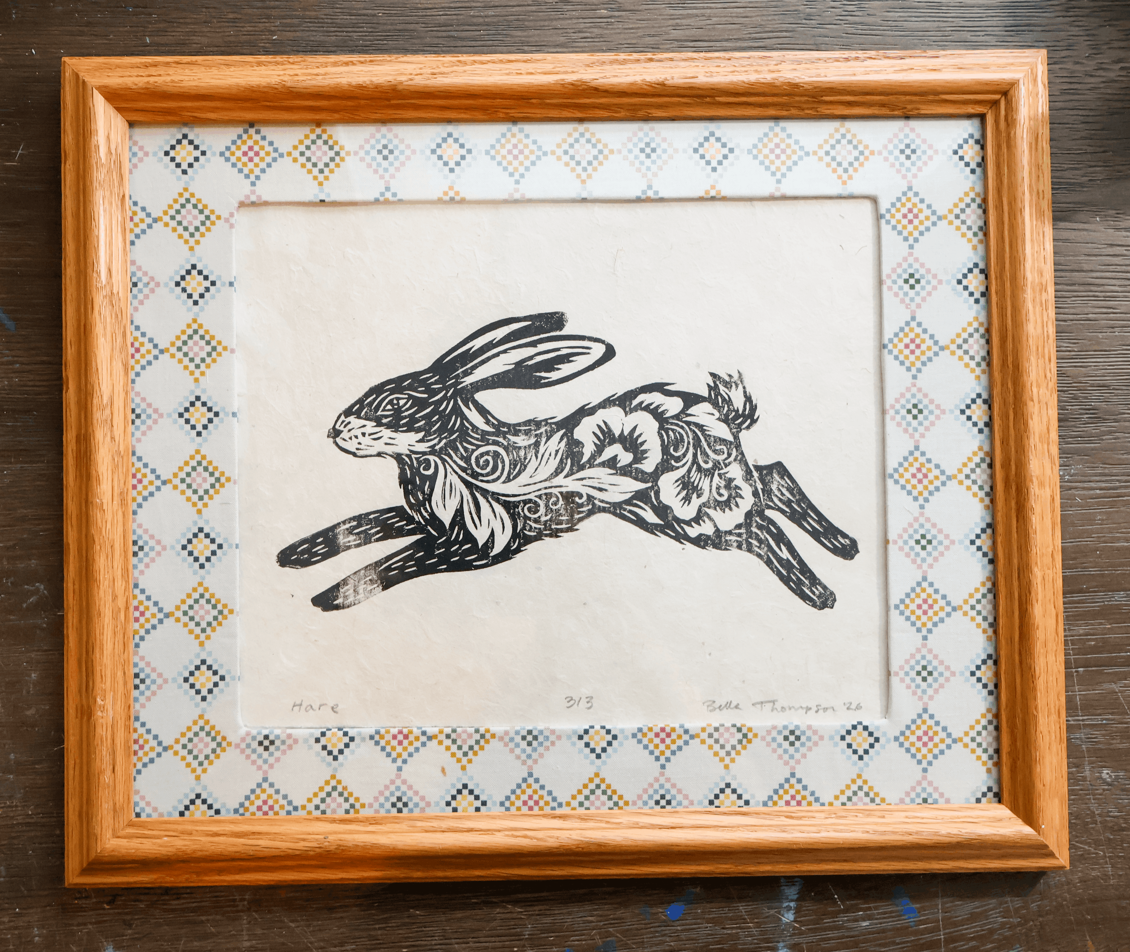 A framed art print featuring a black ink illustration of a leaping rabbit with floral and swirl patterns on its body, set against a cream background with a patterned mat and a light brown wooden frame, displayed on a dark wood surface.