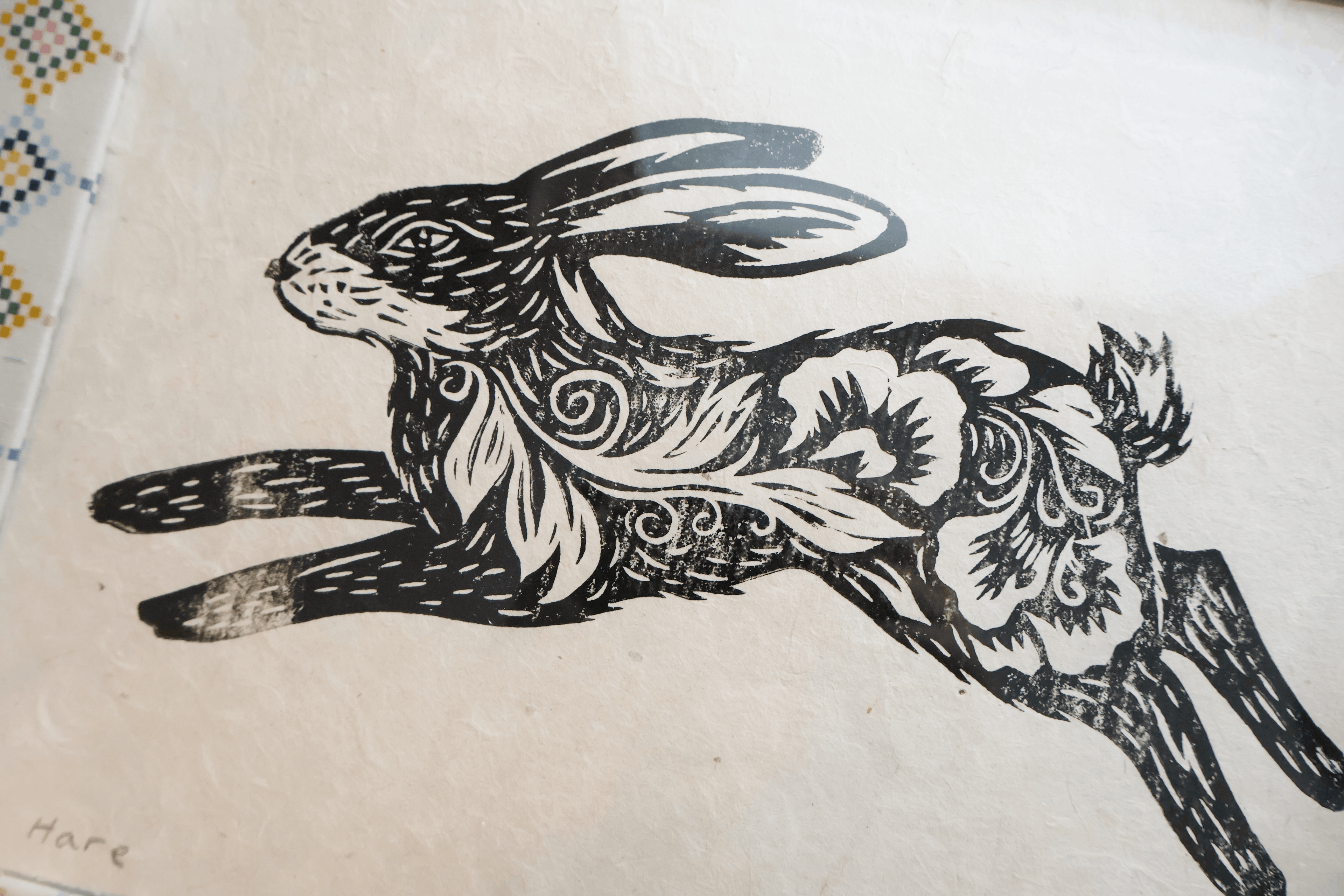 A black ink illustration of a leaping hare with ornate floral and swirl patterns, printed on a textured off-white paper background.