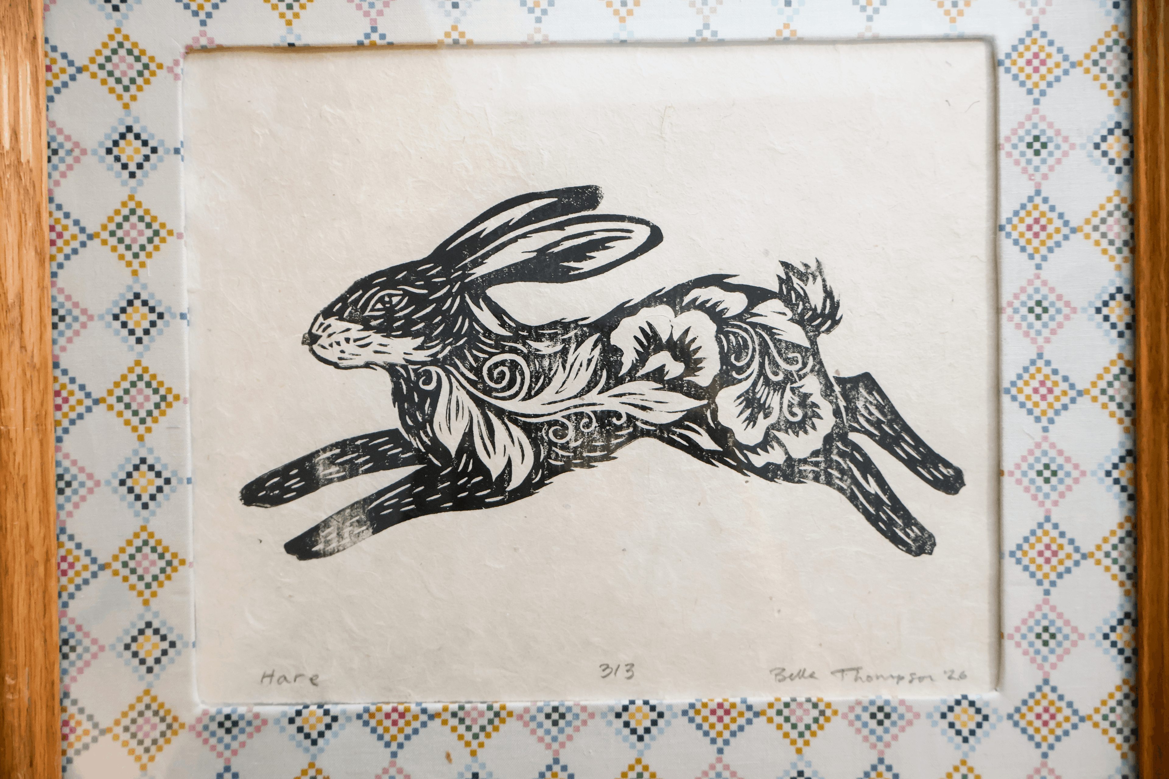 A framed art print featuring a black ink linocut-style rabbit illustration on off-white paper, surrounded by a patterned mat and a light wood frame.