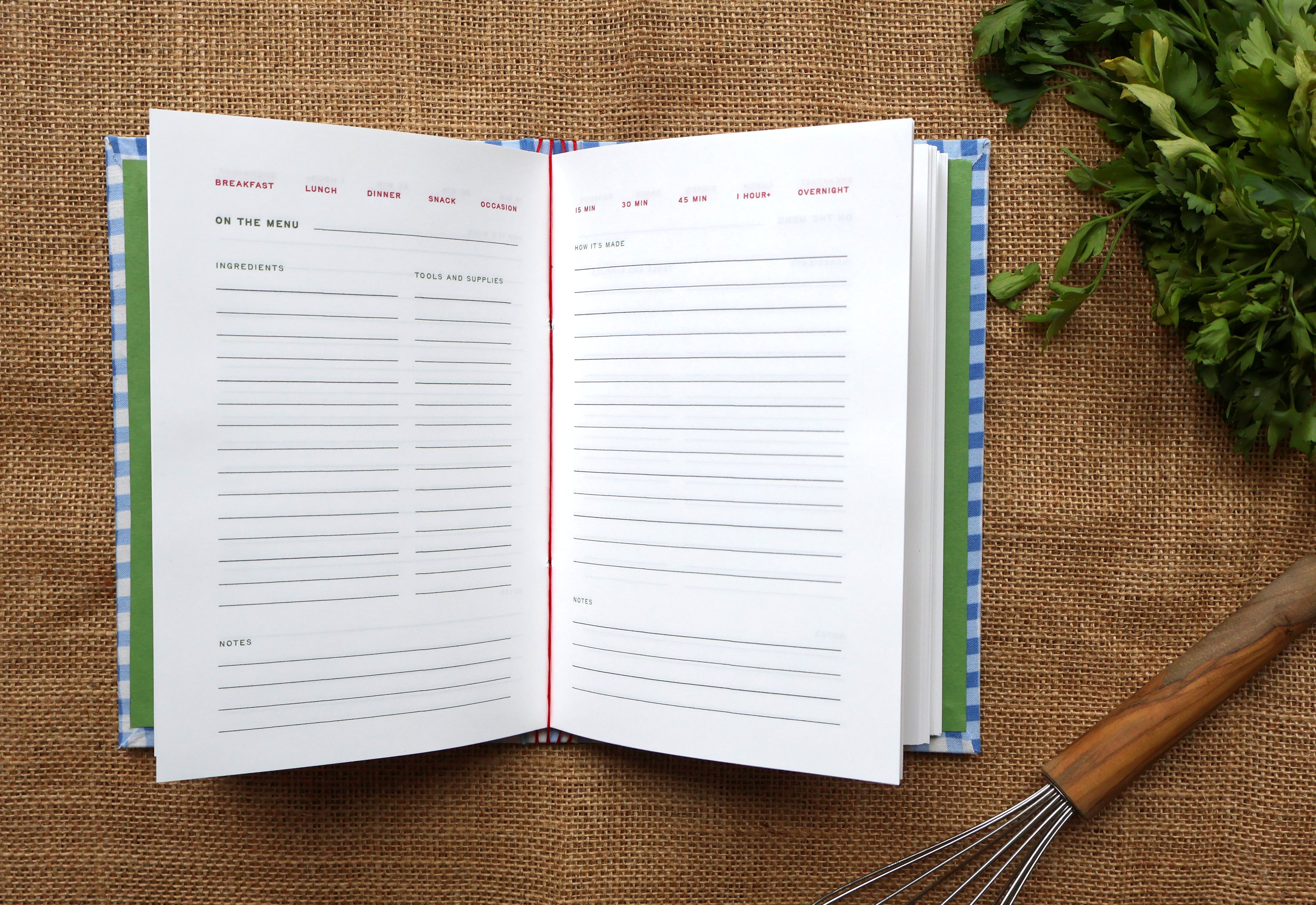 An open recipe journal with lined pages for meals and notes, placed on a burlap surface with green herbs and a wooden-handled whisk nearby.