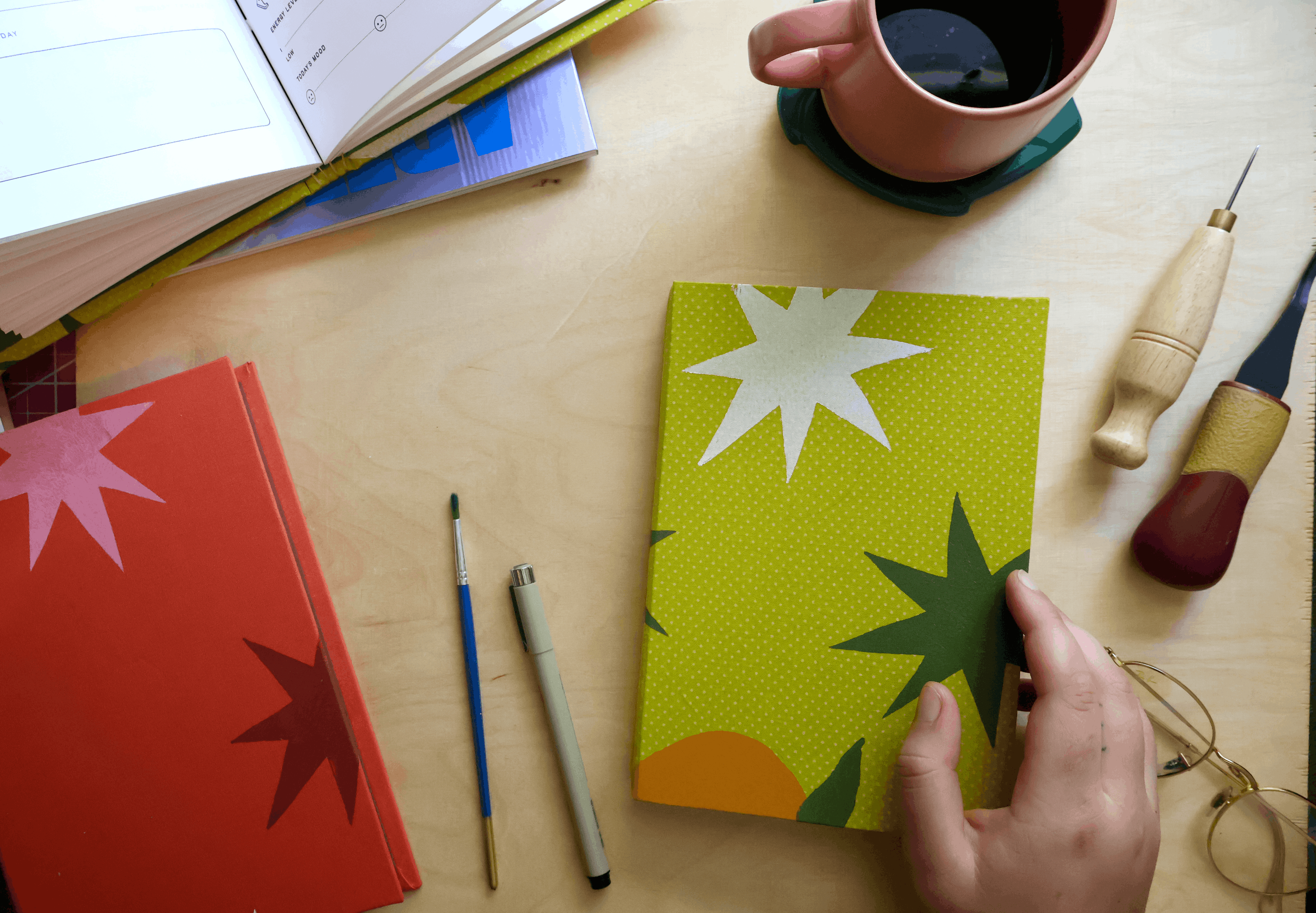 A hand holds a green notebook with a yellow-green textured cover featuring a white star, dark green leaf, and orange sun graphic. The scene is a styled desk flat-lay with other notebooks, a mug, and art tools on a light wood table.