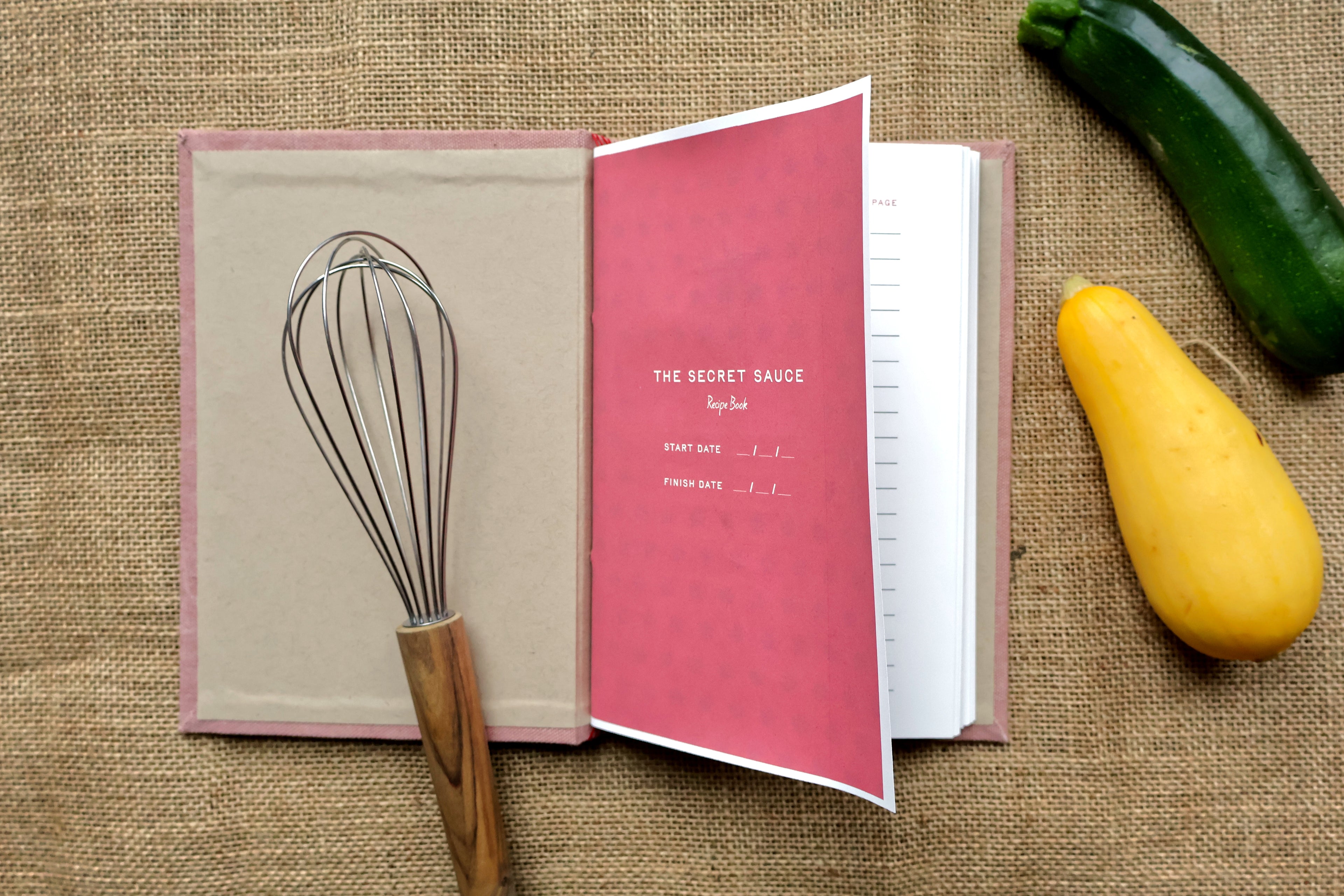 An open recipe log on a burlap surface, with a metal whisk resting on the left page and a pink cover page showing the title 'THE SECRET SAUCE Spyke.' A green zucchini and a yellow squash are placed around it as styled props.