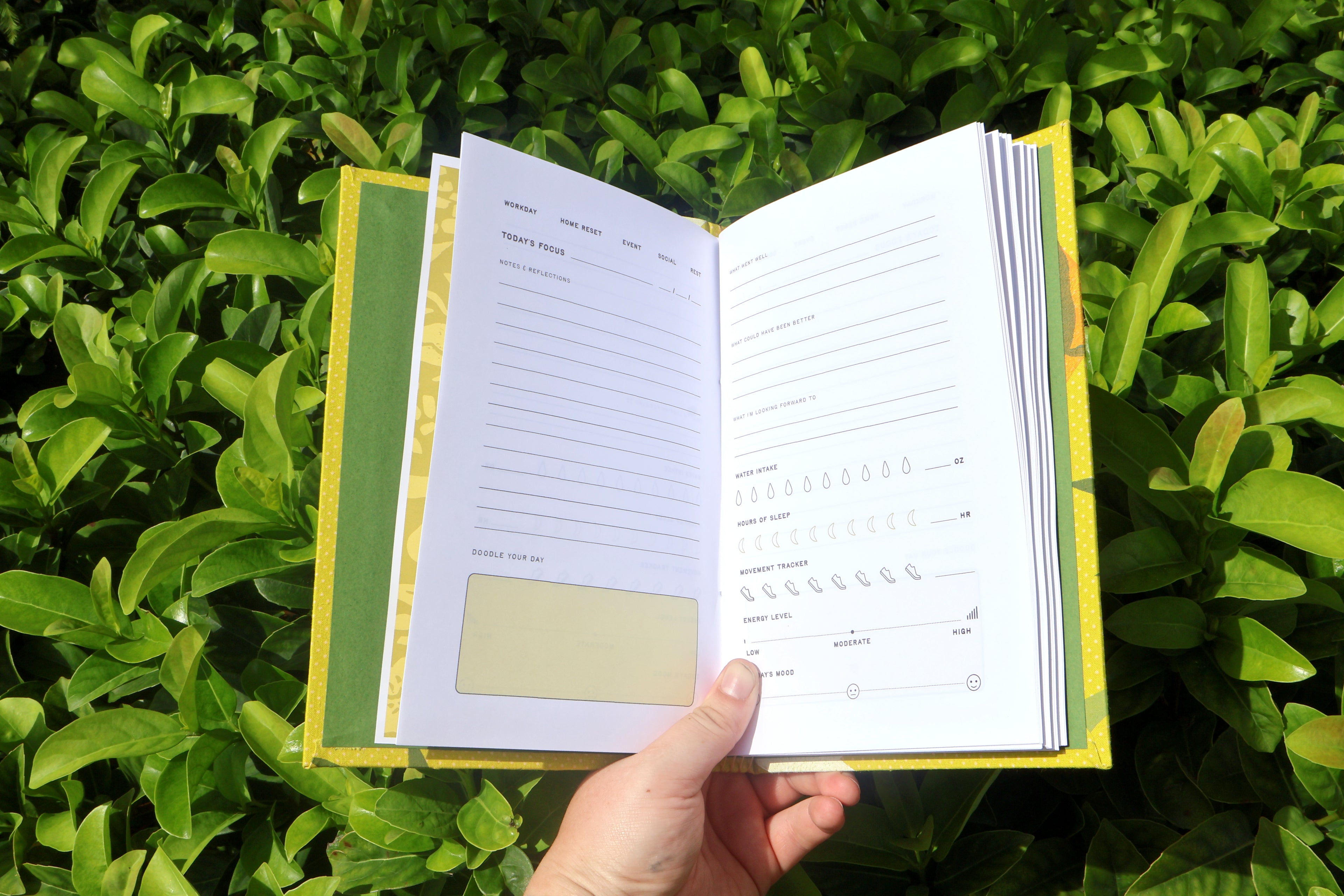 An open notebook or journal with lined pages and a yellow-green cover, held by a hand against a background of green leaves. Hand printed and bound self care reflections log