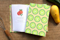 A hardcover recipe log with a green and orange lime slice patterned cover and a white insert page featuring an orange illustration, styled on a burlap surface with a whisk, a yellow squash, and a green zucchini nearby.