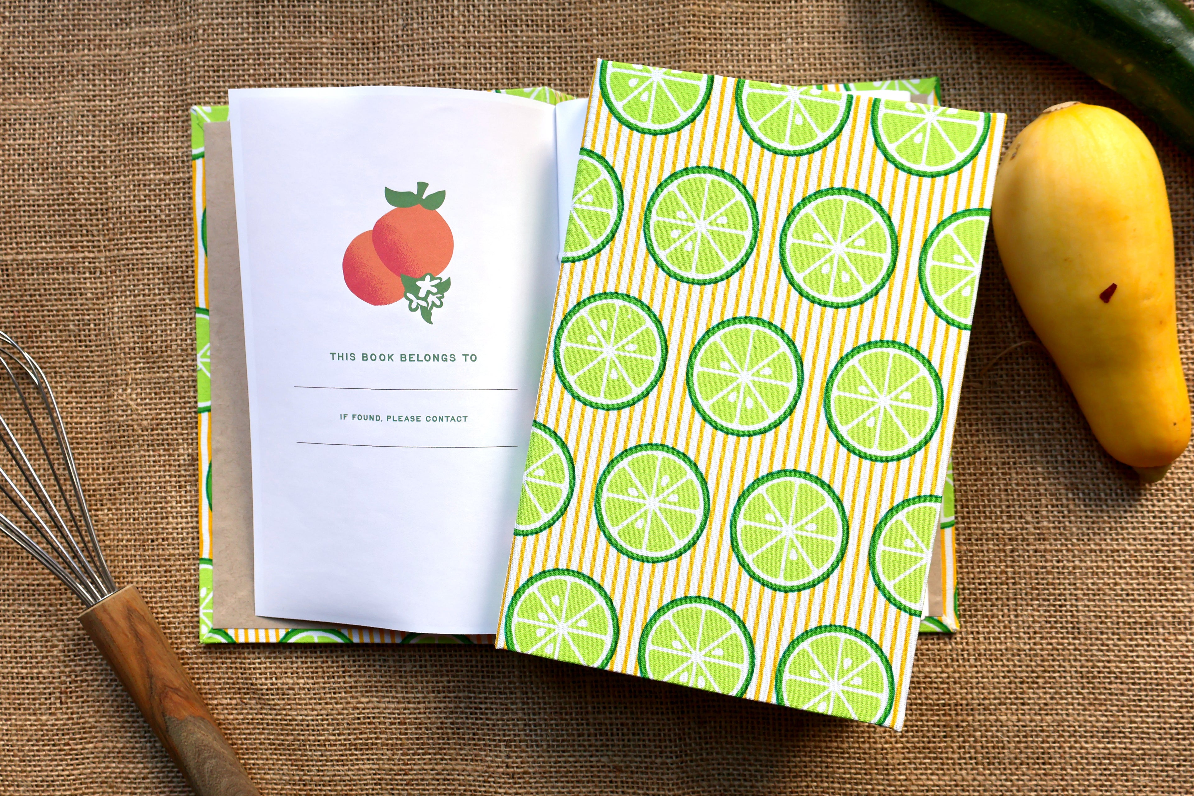A hardcover recipe log with a green and orange lime slice patterned cover and a white insert page featuring an orange illustration, styled on a burlap surface with a whisk, a yellow squash, and a green zucchini nearby.